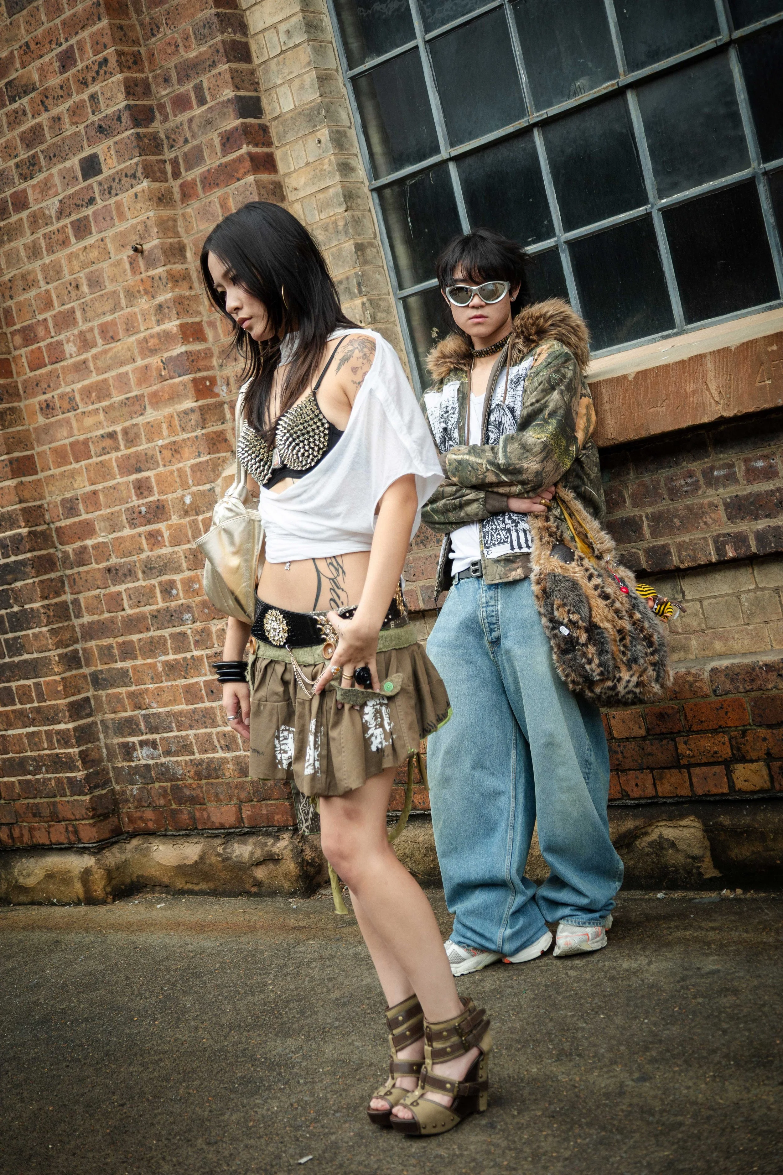 Two young women in punk fashion standing against a brick wall, one with dark hair and studded bra, the other with sunglasses and a furry bag.