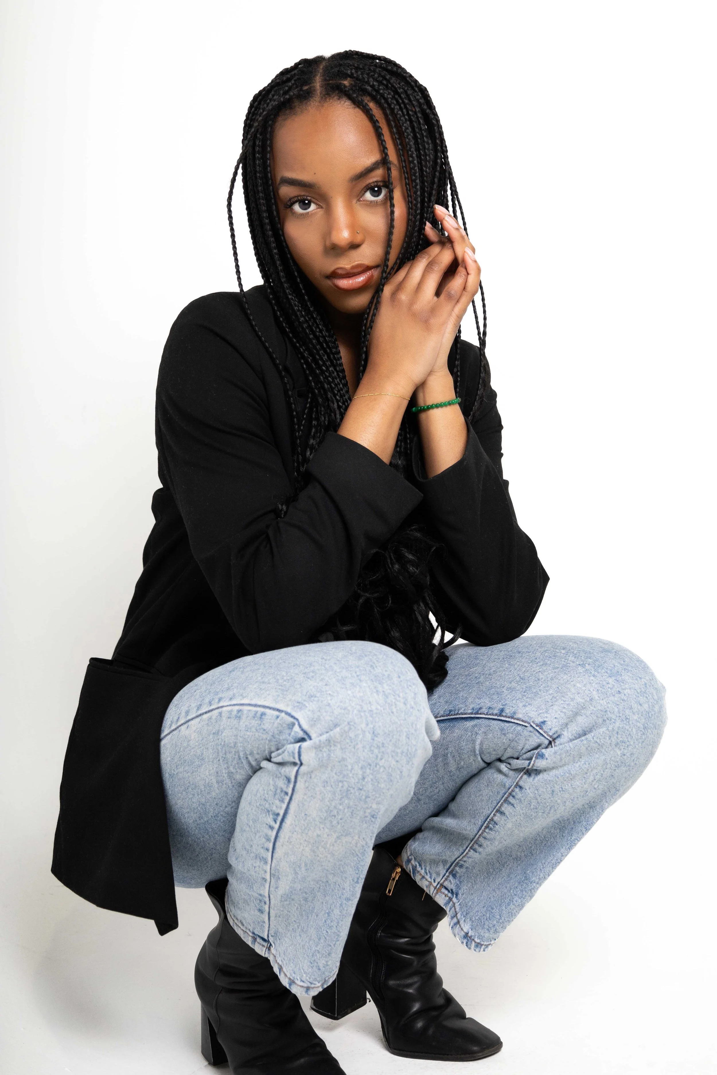 A young woman with braided hair, wearing a black blazer, light blue jeans, and black boots, sitting in a squatting position against a plain white background.