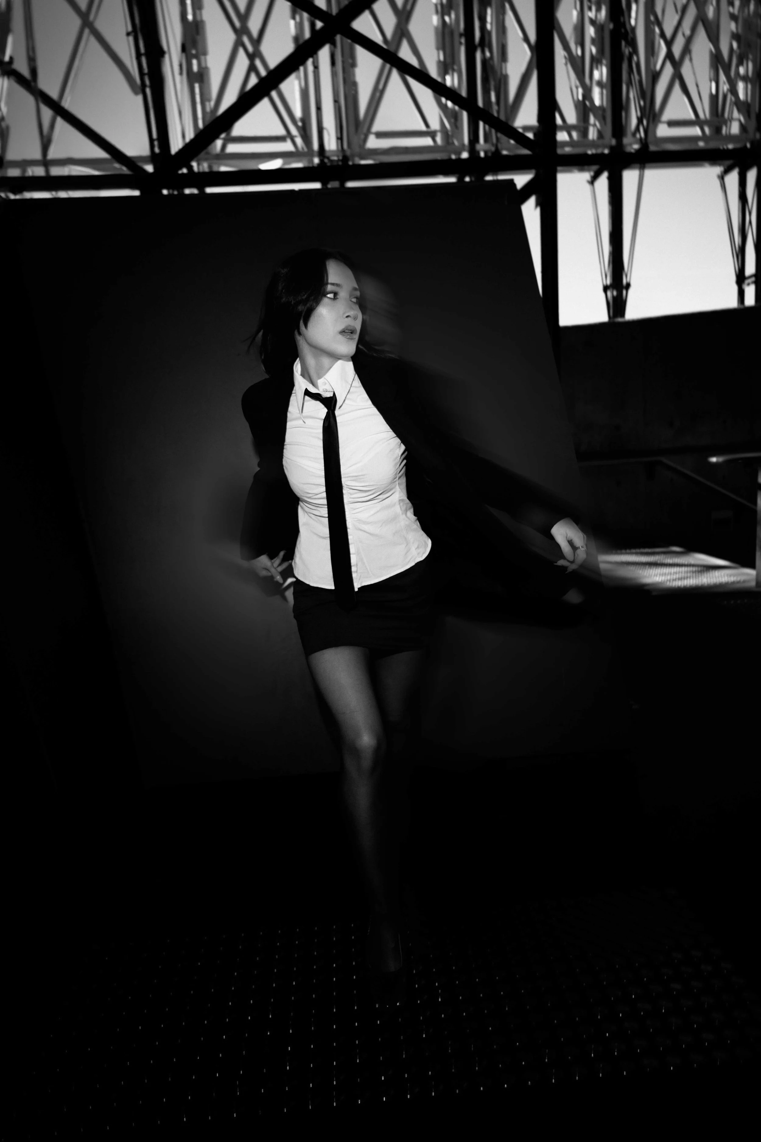 A black and white photo of a woman with dark hair in a business outfit, posing with a dramatic expression against a dark background, with industrial scaffolding and sky above.