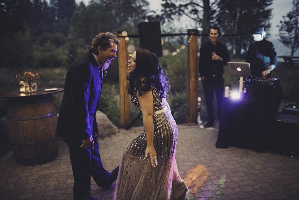Two people dancing and smiling at an outdoor celebration, with two musicians in the background, one with a laptop and DJ equipment, at dusk with string lights overhead.