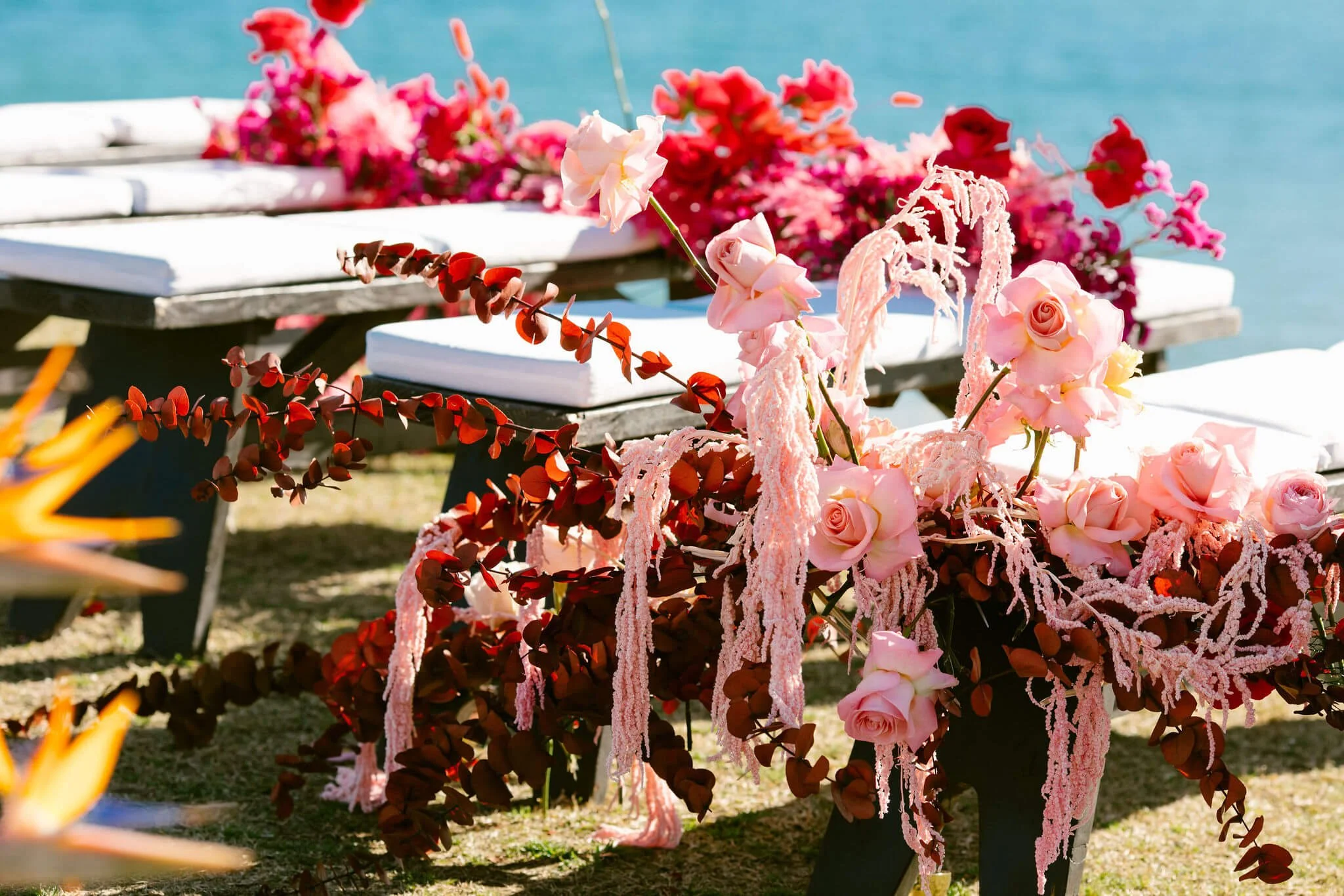 Colorful floral arrangements with pink roses, red and pink flowers, and ribbons on wooden tables near a body of water.