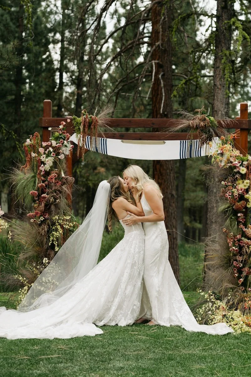 Two brides in wedding dresses share a kiss under a wooden arch decorated with flowers and greenery in a forest setting.