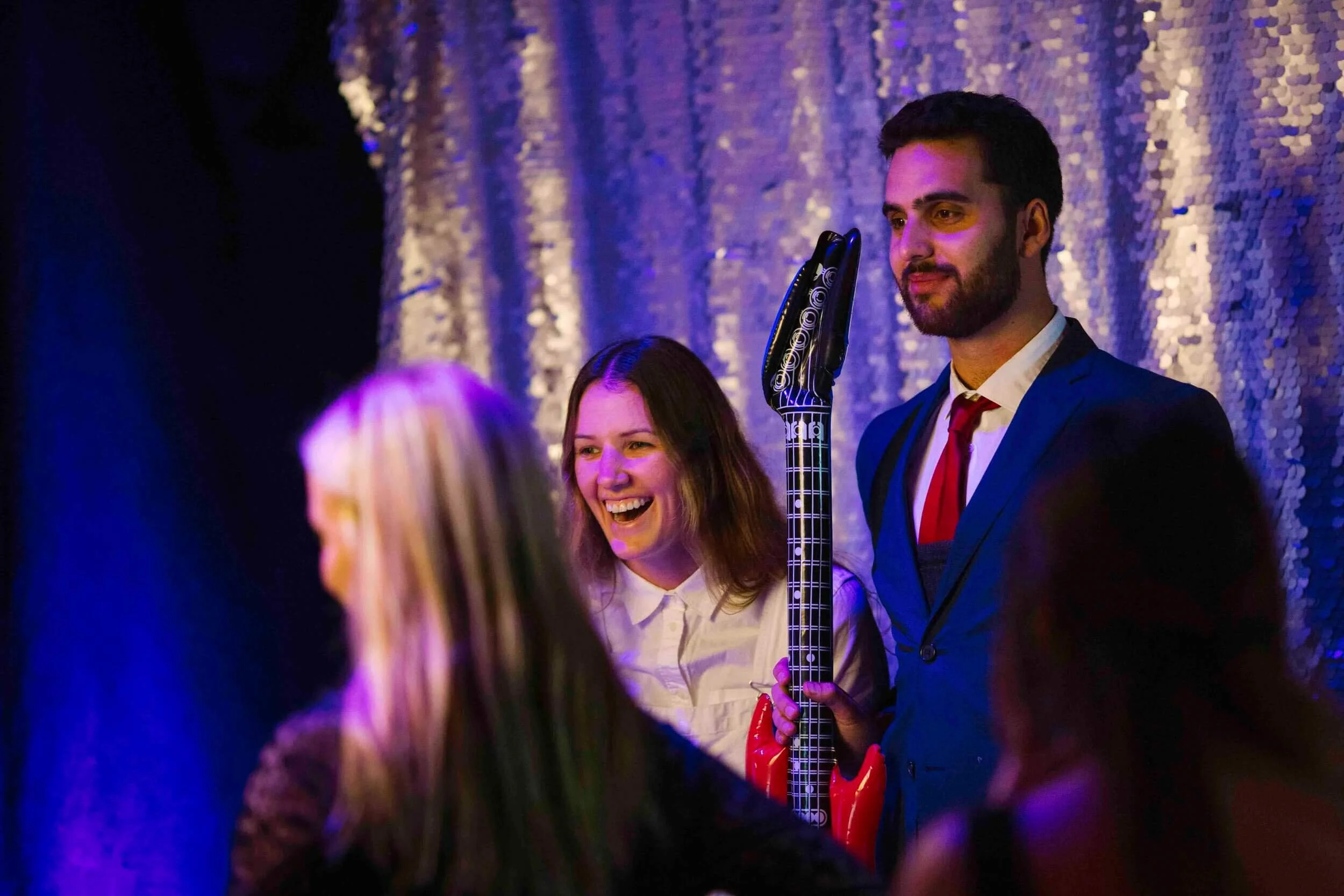 A man in a blue suit and red tie, holding a guitar-shaped device, standing next to a smiling woman in a white shirt at a party or event with a sequined backdrop