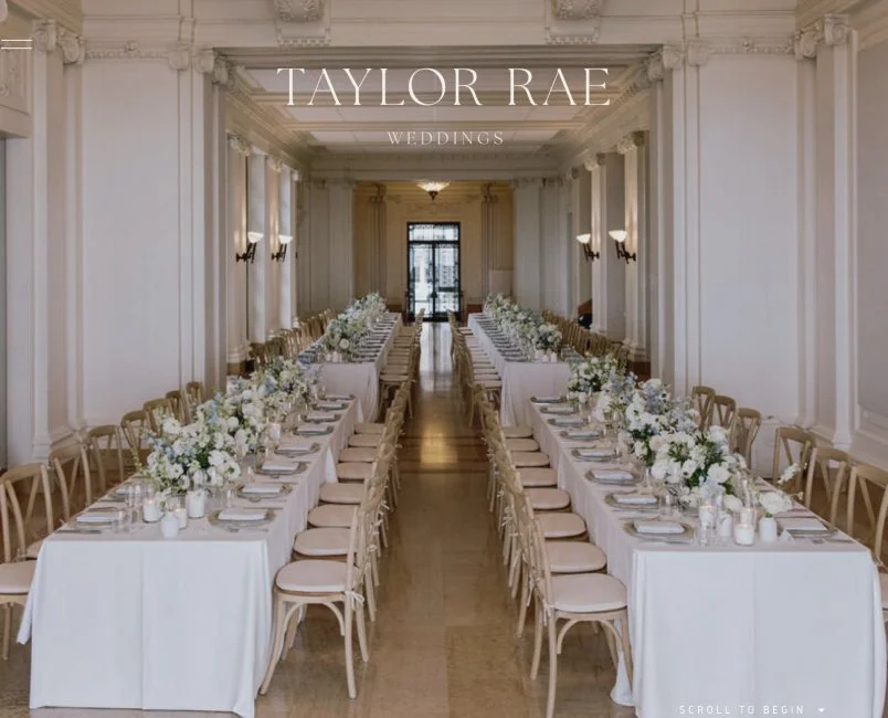 Elegant wedding reception setup with long banquet tables decorated with white floral centerpieces, set with plates and silverware, in a grand, historic hall with high ceiling, ornate molding, and large windows.
