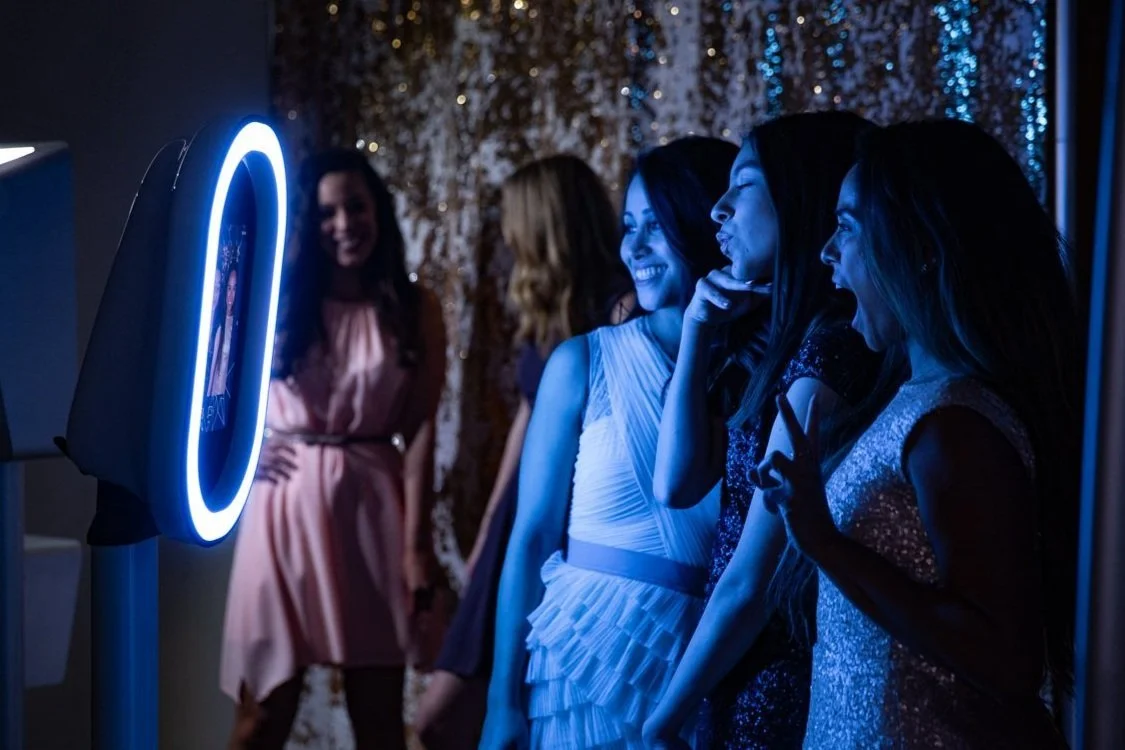 Four women laughing and taking pictures at a photo booth with a dark background and bright blue lighting.
