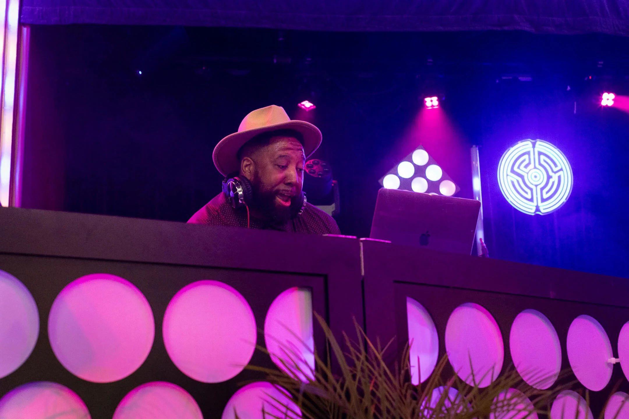 Man DJ wearing a beige hat and headphones, using a laptop, with purple and pink stage lighting in the background.