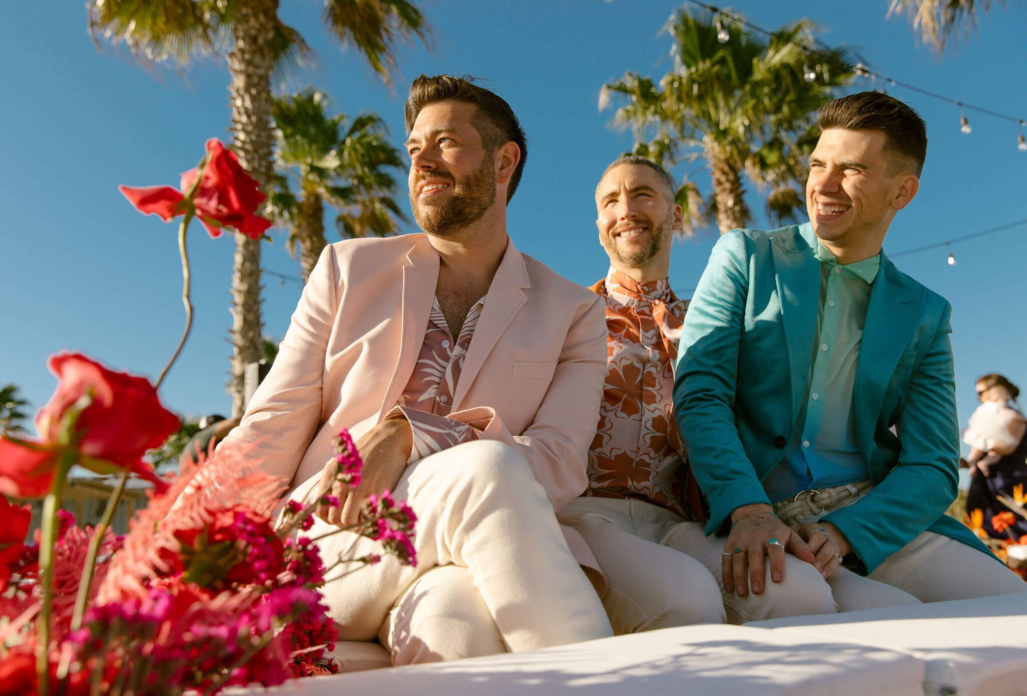 Three men in colorful suits sitting outdoors during a sunny day with palm trees, flowers, and string lights in the background.