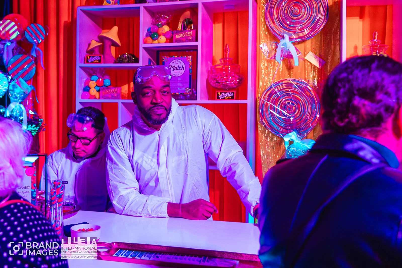 People playing a colorful arcade game with neon lights and shelves filled with toys and candies in the background.