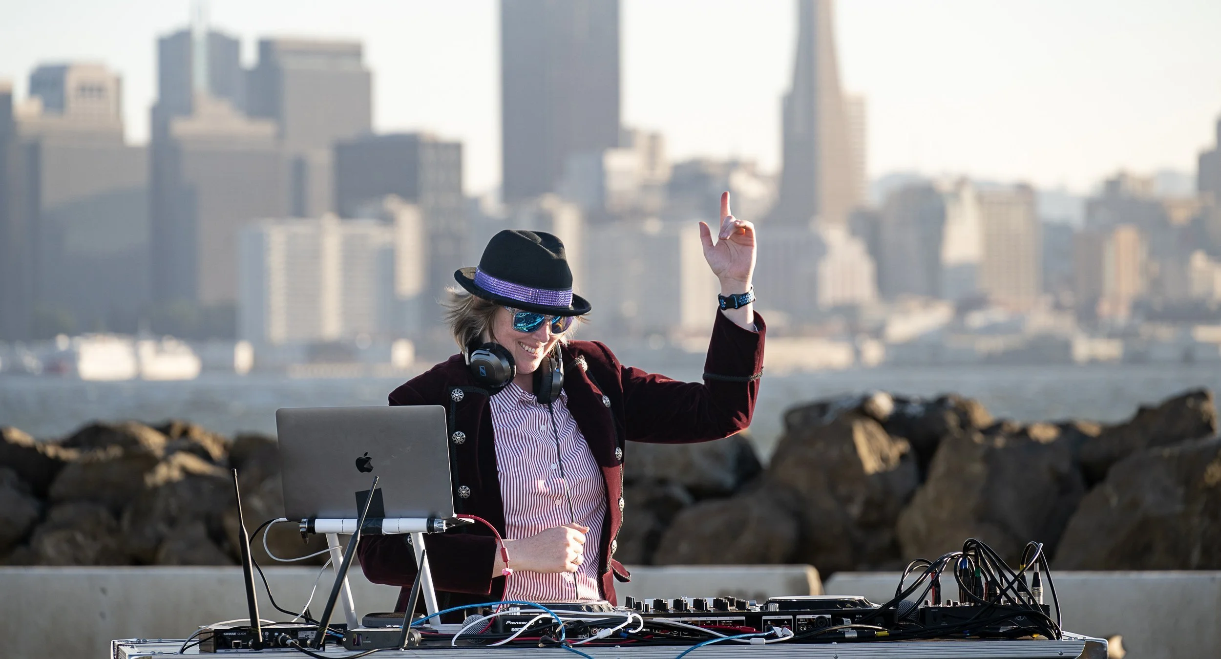 A female DJ wearing sunglasses, a hat, and headphones is performing outdoors near a body of water with a city skyline in the background. She is smiling, raising one arm, and using DJ equipment on a table.