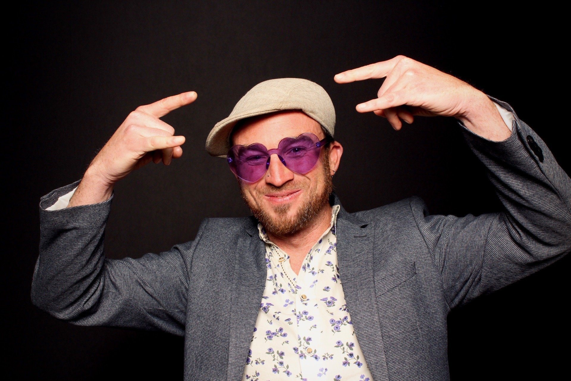 Man wearing purple heart-shaped sunglasses, a beige beret, a patterned shirt, and a gray blazer, smiling and pointing to his head with both hands, against a black background.