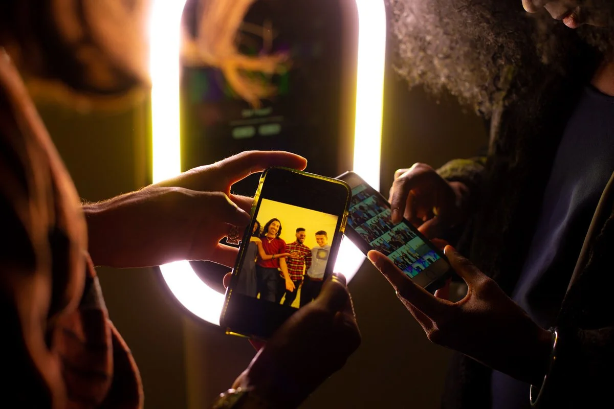 Two people taking a selfie with a smartphone, the background features a bright ring light and another person using a smartphone to view photo thumbnails.