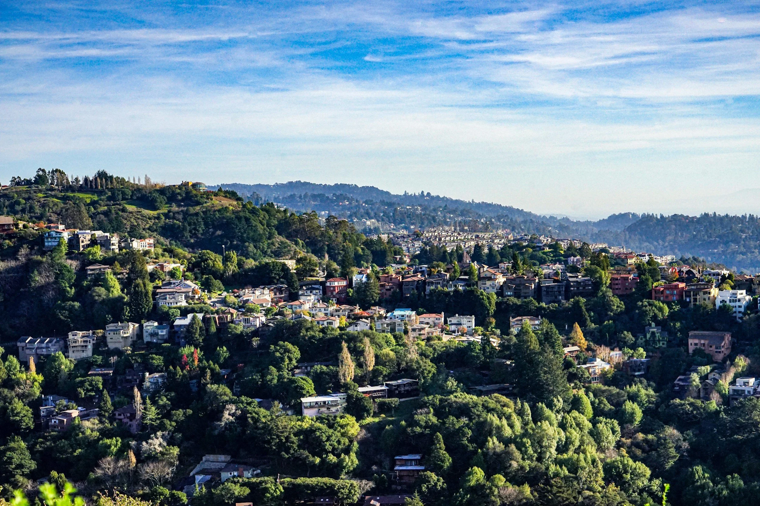 Hilly landscape with numerous houses and green trees under a blue sky with wispy clouds.