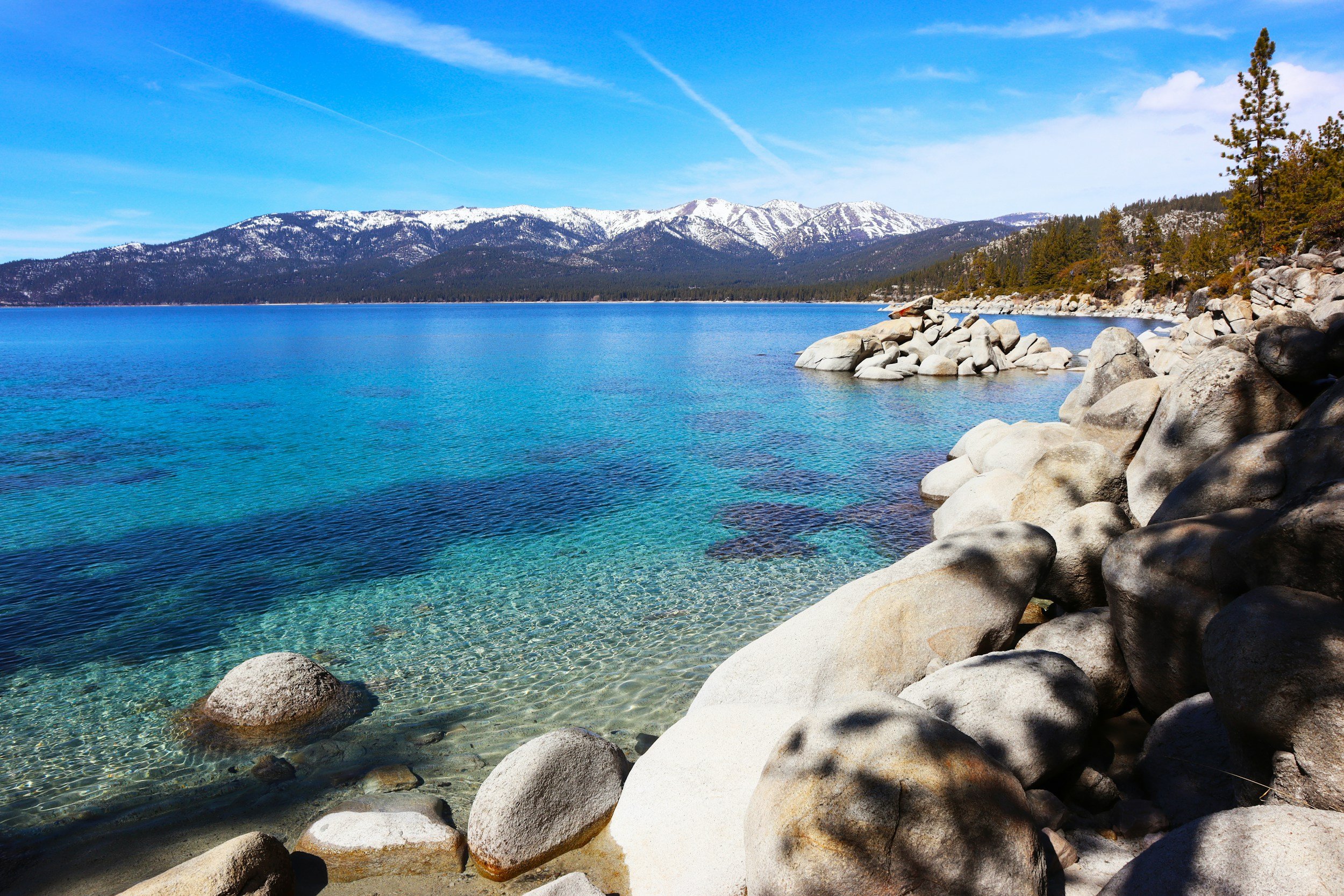 A scenic view of a clear blue lake with a rocky shoreline, pine trees, and snow-capped mountains in the background under a blue sky with a few clouds.