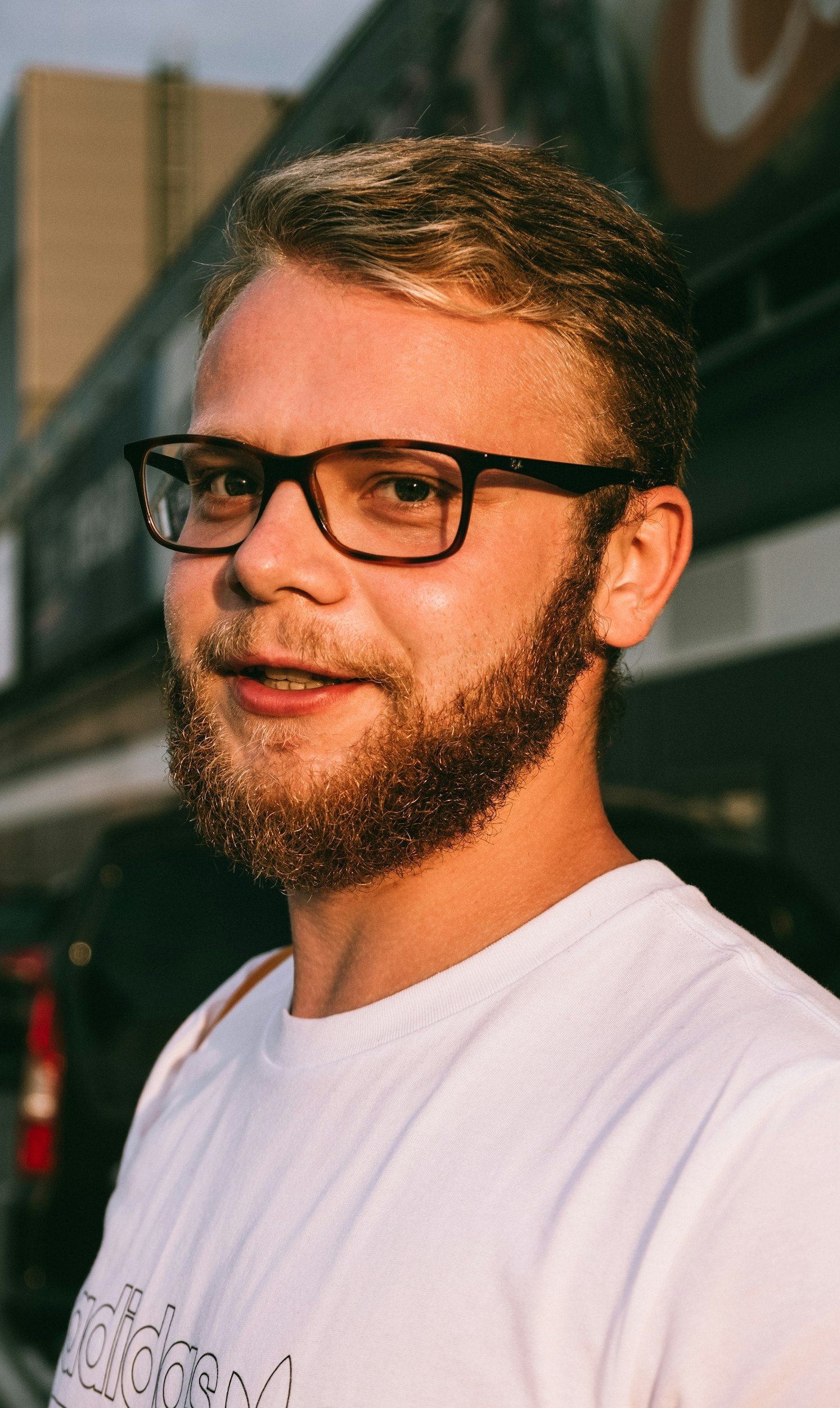 A young man with a beard wearing glasses and a white t-shirt, smiling outdoors in front of a dark vehicle and building, during sunset.