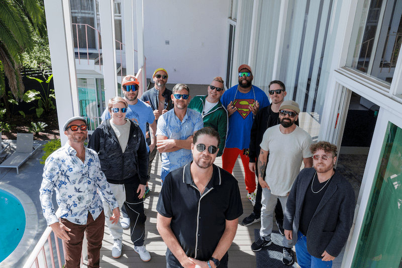 A group of ten men standing on a balcony, dressed casually with some wearing sunglasses and hats, posing for a photo with a pool and modern building in the background.