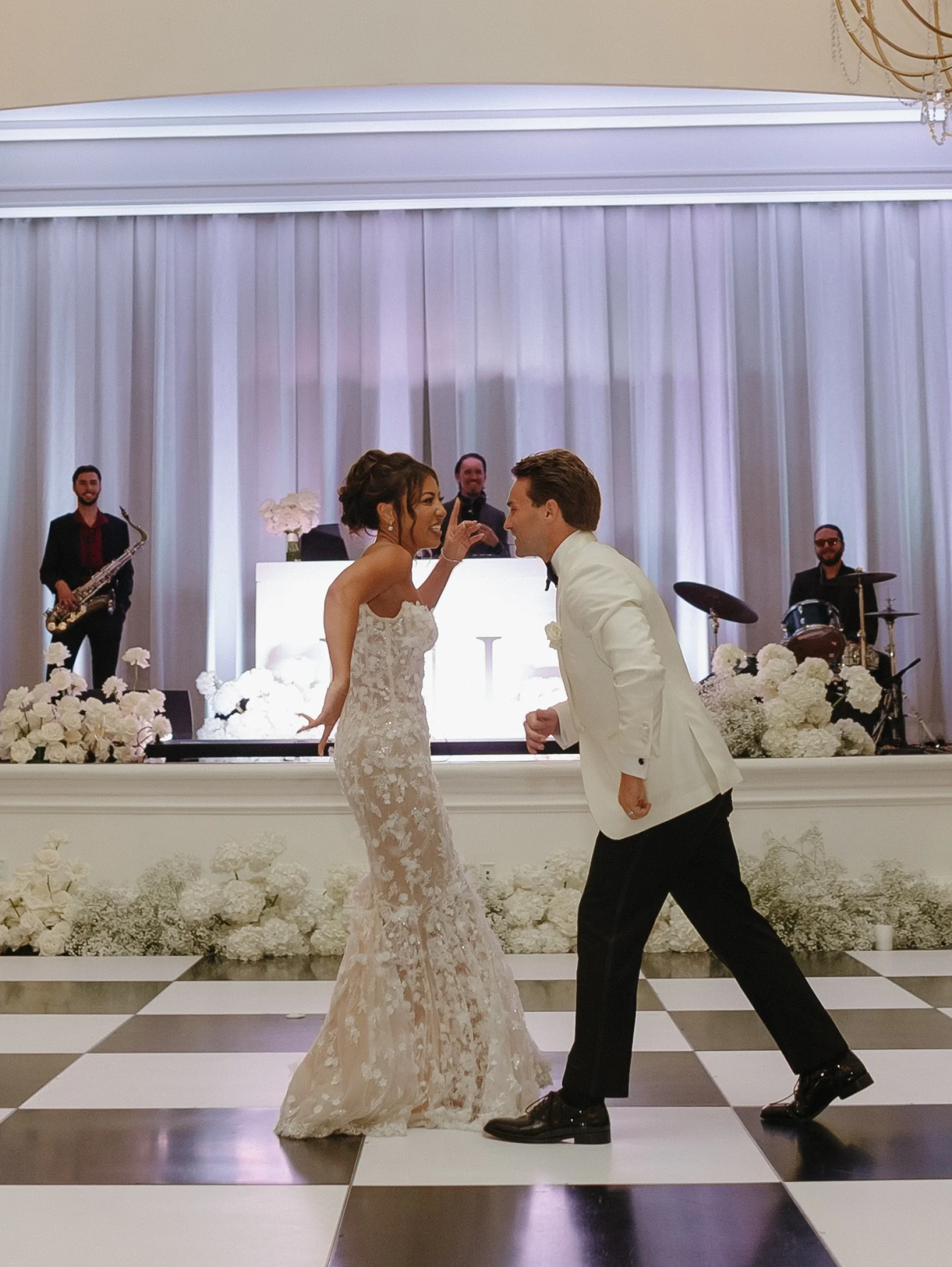 A bride and groom dancing together on a checkered dance floor at their wedding reception, with a live band playing in the background.