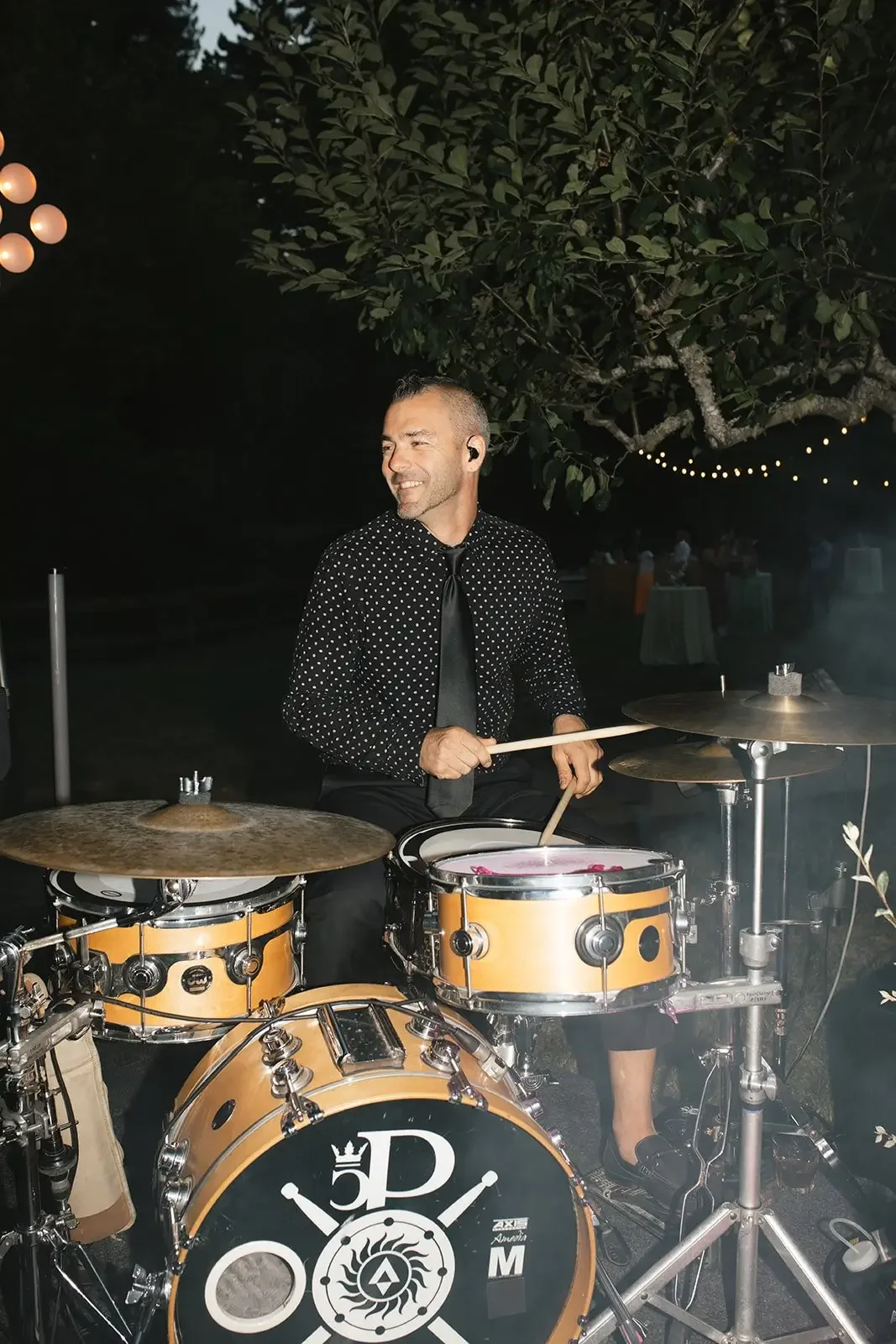 A man playing drums outdoors at night, dressed in a black shirt with white polka dots and a black tie, with string lights and a tree in the background.