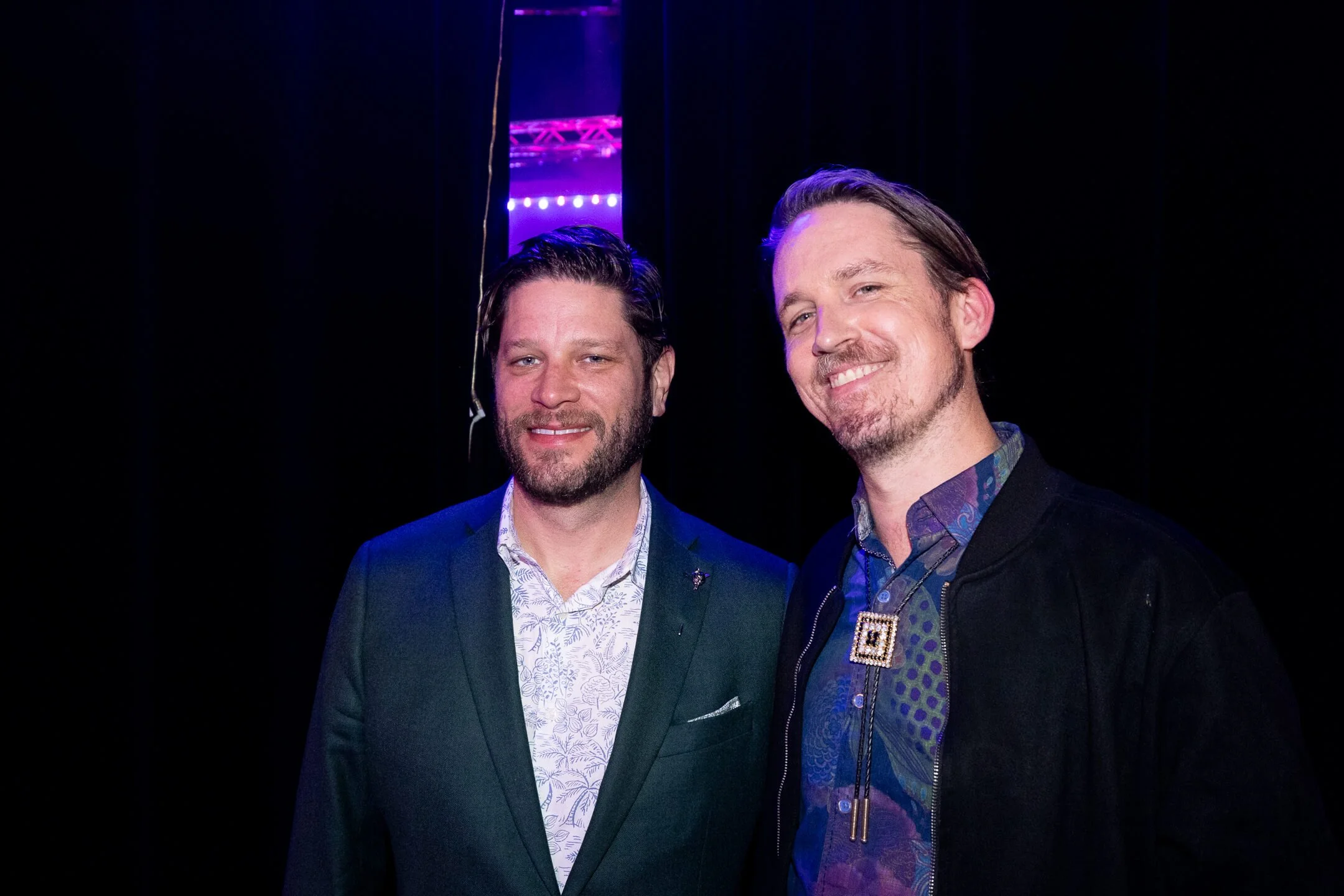 Two men smiling, standing in front of a dark background with purple and blue lighting.