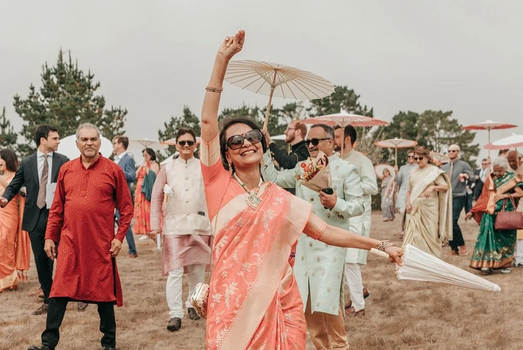 A woman in a pink sari holding an umbrella and smiling at an outdoor celebration with other people dressed in traditional Indian attire, some holding umbrellas.