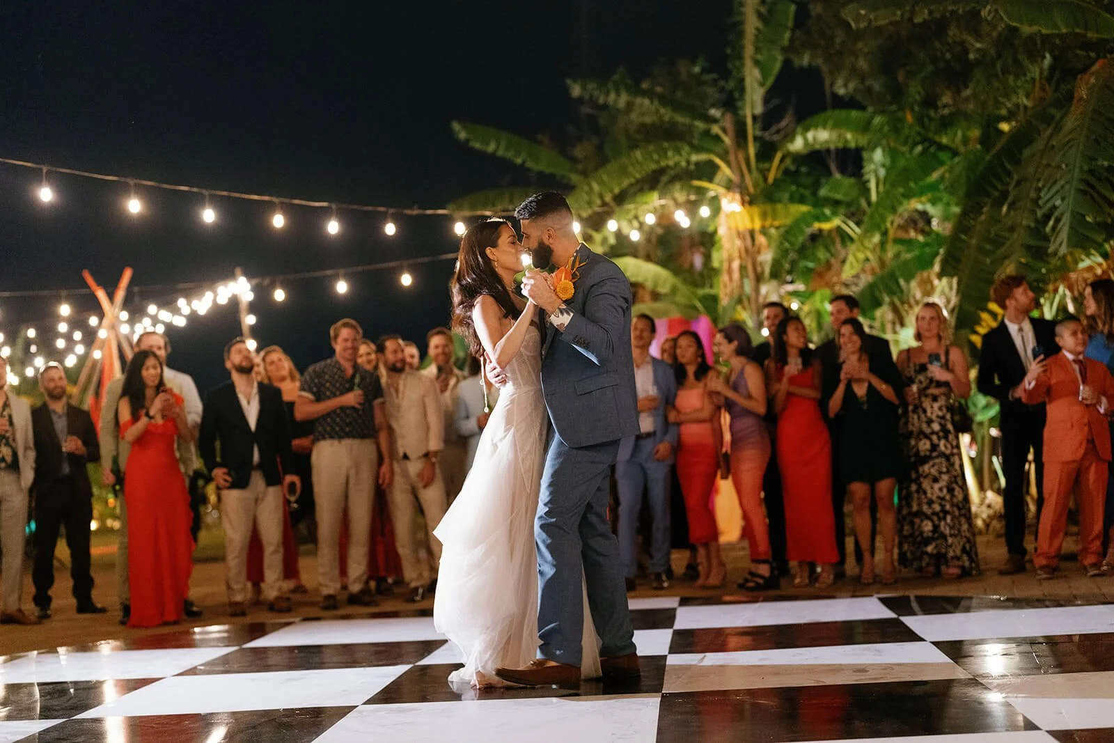 A couple's first dance at their wedding reception, surrounded by friends and family, outdoors at night with string lights and tropical plants in the background.