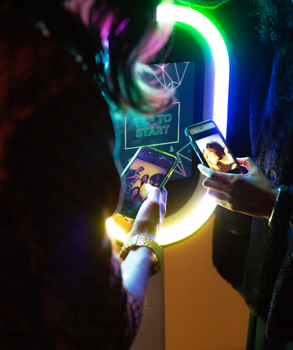 Two people taking photos of each other with smartphones in front of a colorful, neon-lit photo booth or display with a sign that says 'Tap to Start'.