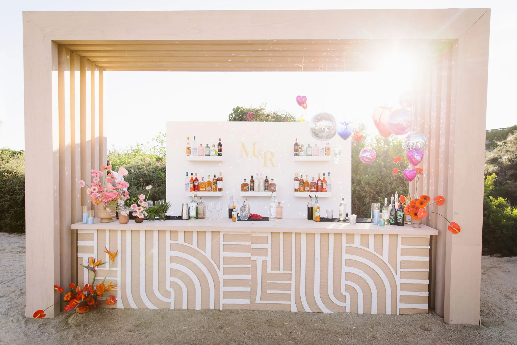 Decorated outdoor bar with bottles of alcohol, pink and orange flowers, floating heart-shaped balloons, and a white backdrop with gold initials 'M & R' under a wooden frame