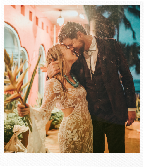 A couple, a woman in a lace wedding dress and a man in a suit, share a romantic moment in the rain, smiling and touching foreheads outside at night with a pink building and tropical plants in the background.
