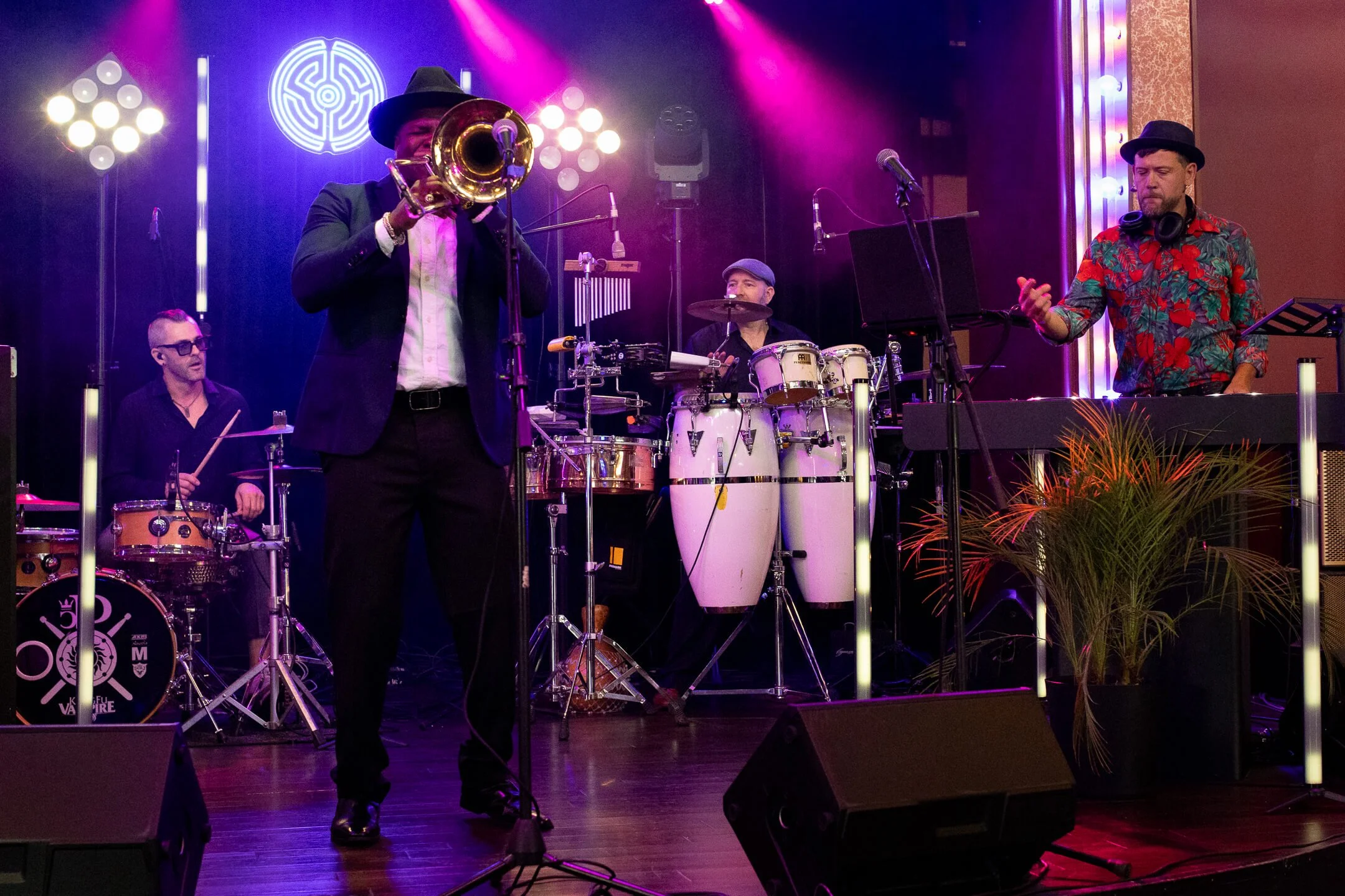 A band performing live on stage with colorful lighting. One musician is playing a trumpet, another on drums, a third on congas, and a fourth on a keyboard. The drummer and keyboardist are wearing hats, and the band is accompanied by stage lights and a potted plant.