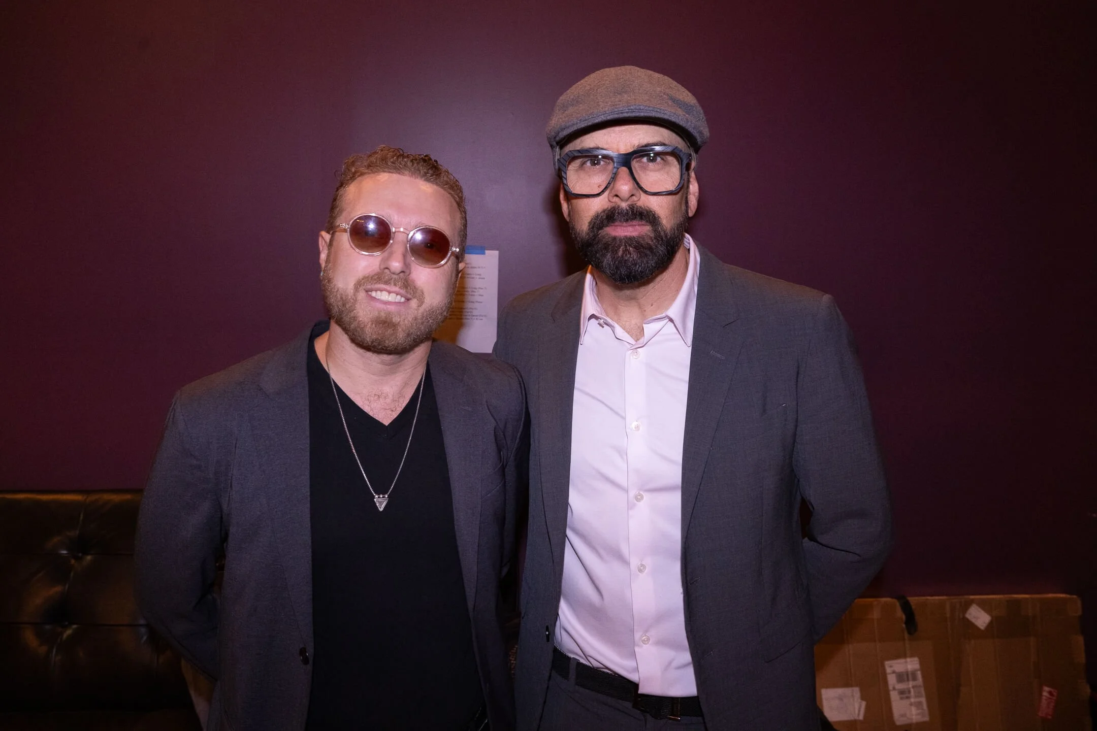 Two men standing together in a dimly lit room, wearing suits. The man on the left has curly hair, a beard, and is wearing sunglasses, a necklace, and a dark blazer. The man on the right has a beard, glasses, a flat cap, and a light-colored shirt unde