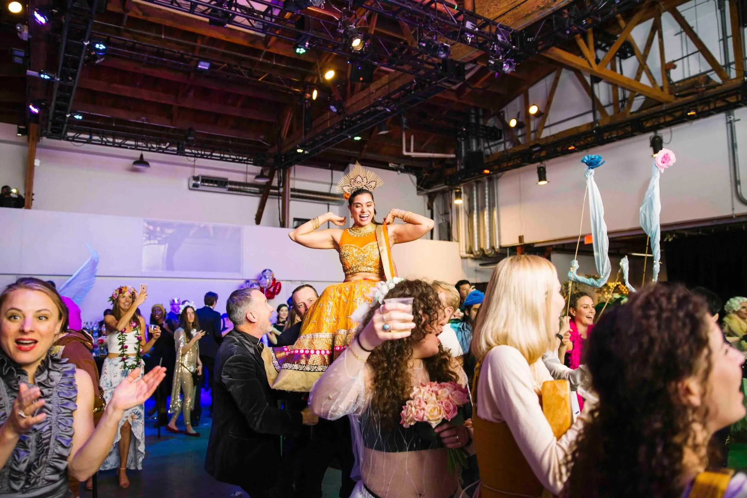 A group of people celebrating at a party or event in an indoor venue with a high wooden ceiling. A woman dressed in a bright yellow traditional outfit with a decorative crown is standing on a chair, smiling, with arms flexed. Others around are clappi