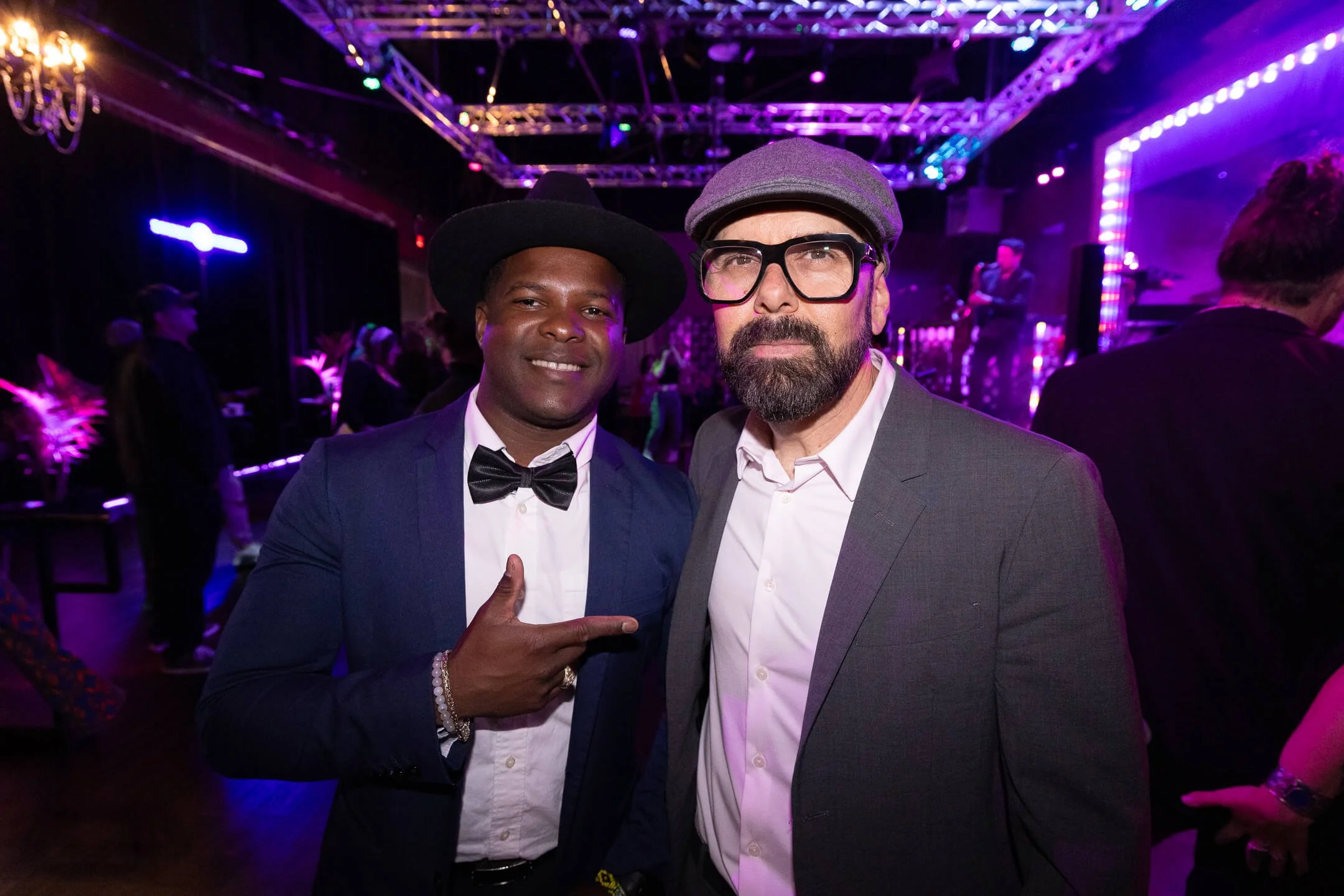 Two men dressed in suits at a nightclub or party with colorful purple and blue lighting, one pointing at the other.