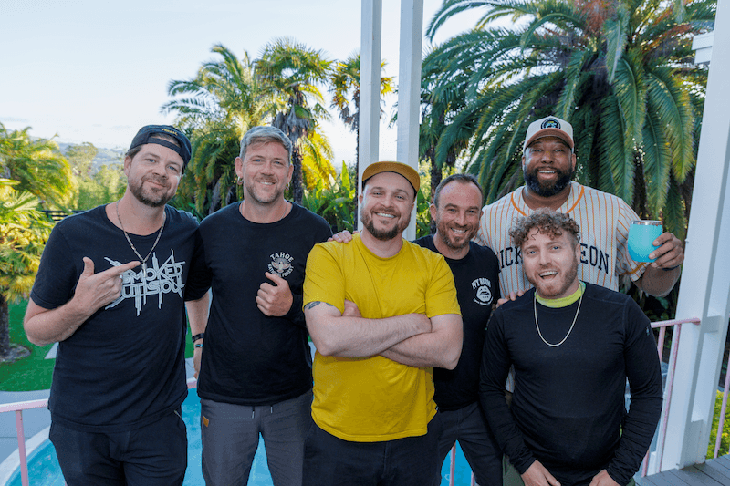 Group of six men smiling and posing on a balcony with lush tropical plants in the background, some holding drinks.