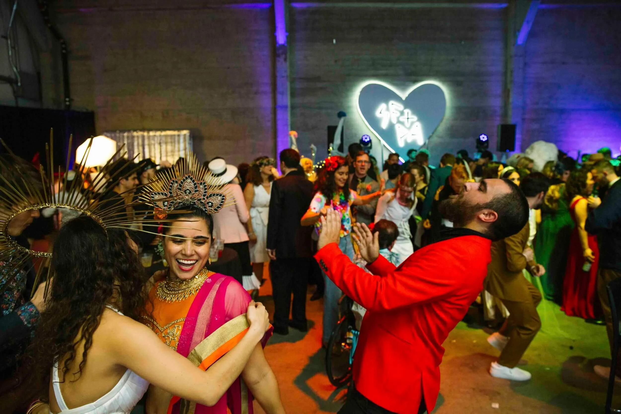 People dancing and celebrating at a colorful party with a neon heart and Chinese characters on the wall in the background.