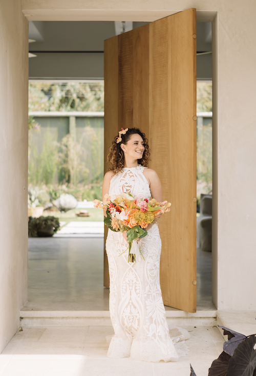 A woman in a white lace dress holding a bouquet of colorful flowers, standing in a doorway and smiling.