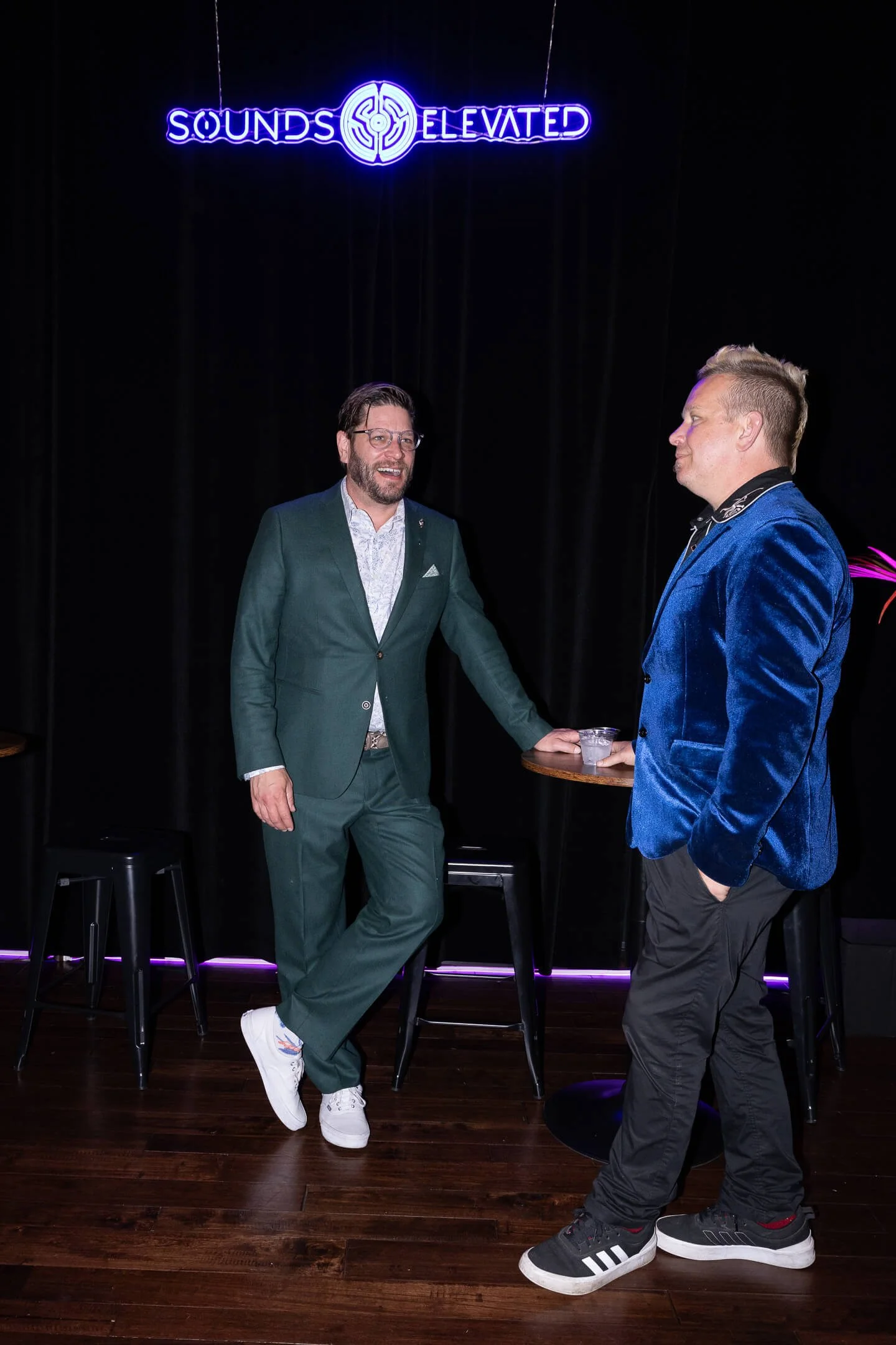 Two men in suits talking at a bar, with a neon sign above them that reads 'Sounds Elevated.'
