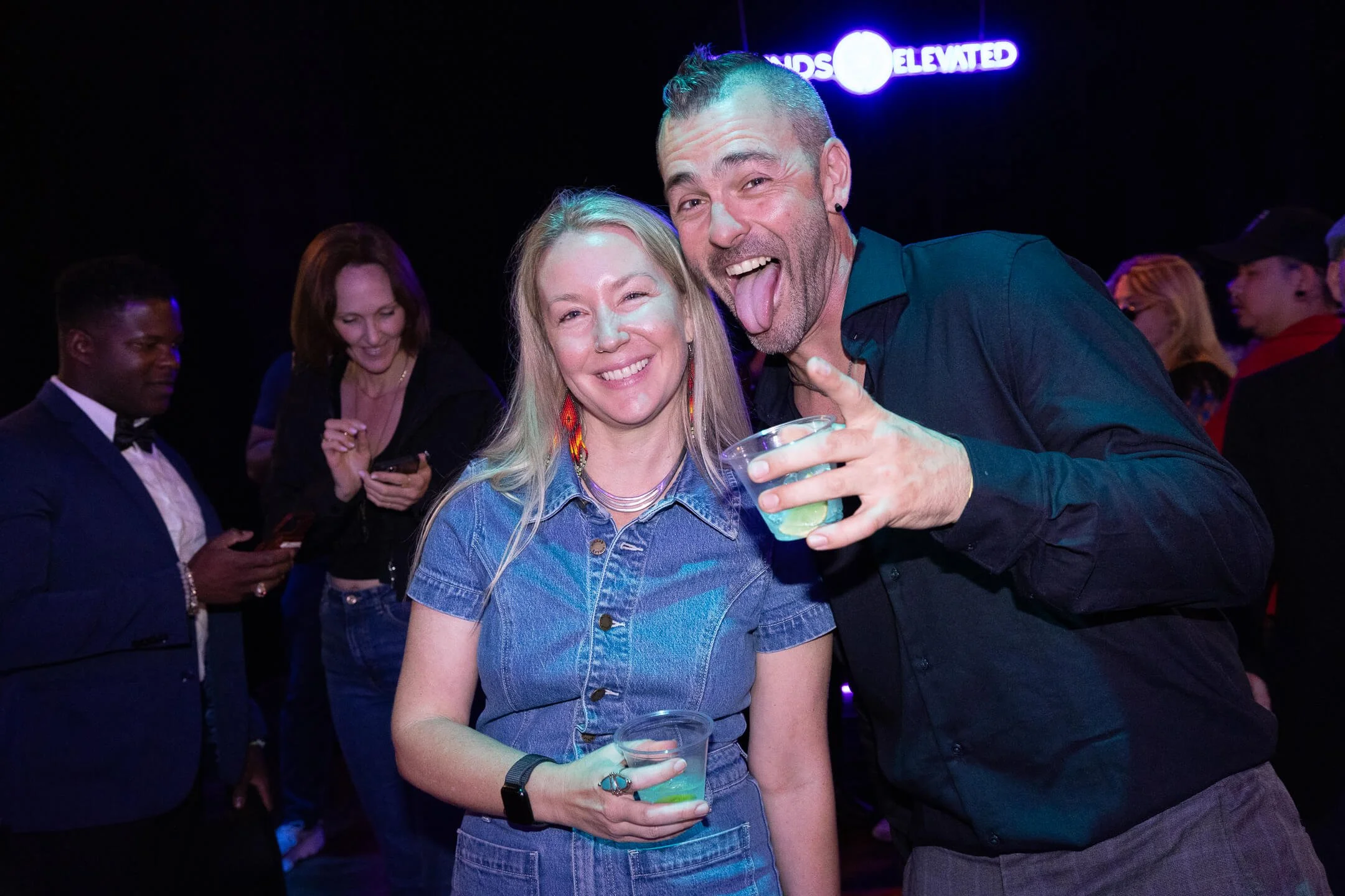 Two friends smiling and posing for a photo at a party or nightclub. The woman is holding a drink and wearing a denim dress with colorful earrings, while the man is making a funny face with his mouth open and tongue out, holding a drink. Background fe