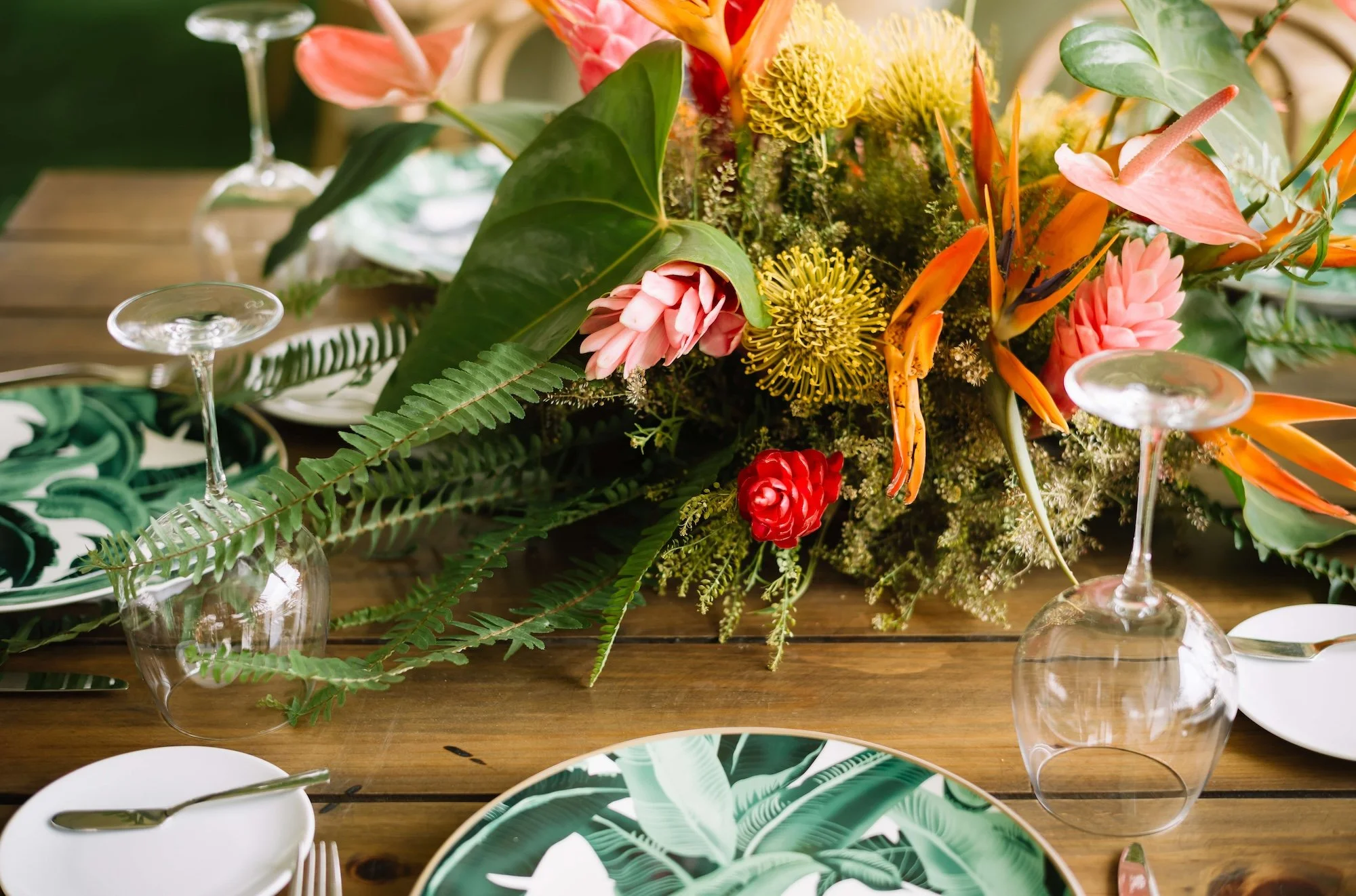 A table setting with a floral centerpiece, tropical-themed plates, and glassware on a wooden table.
