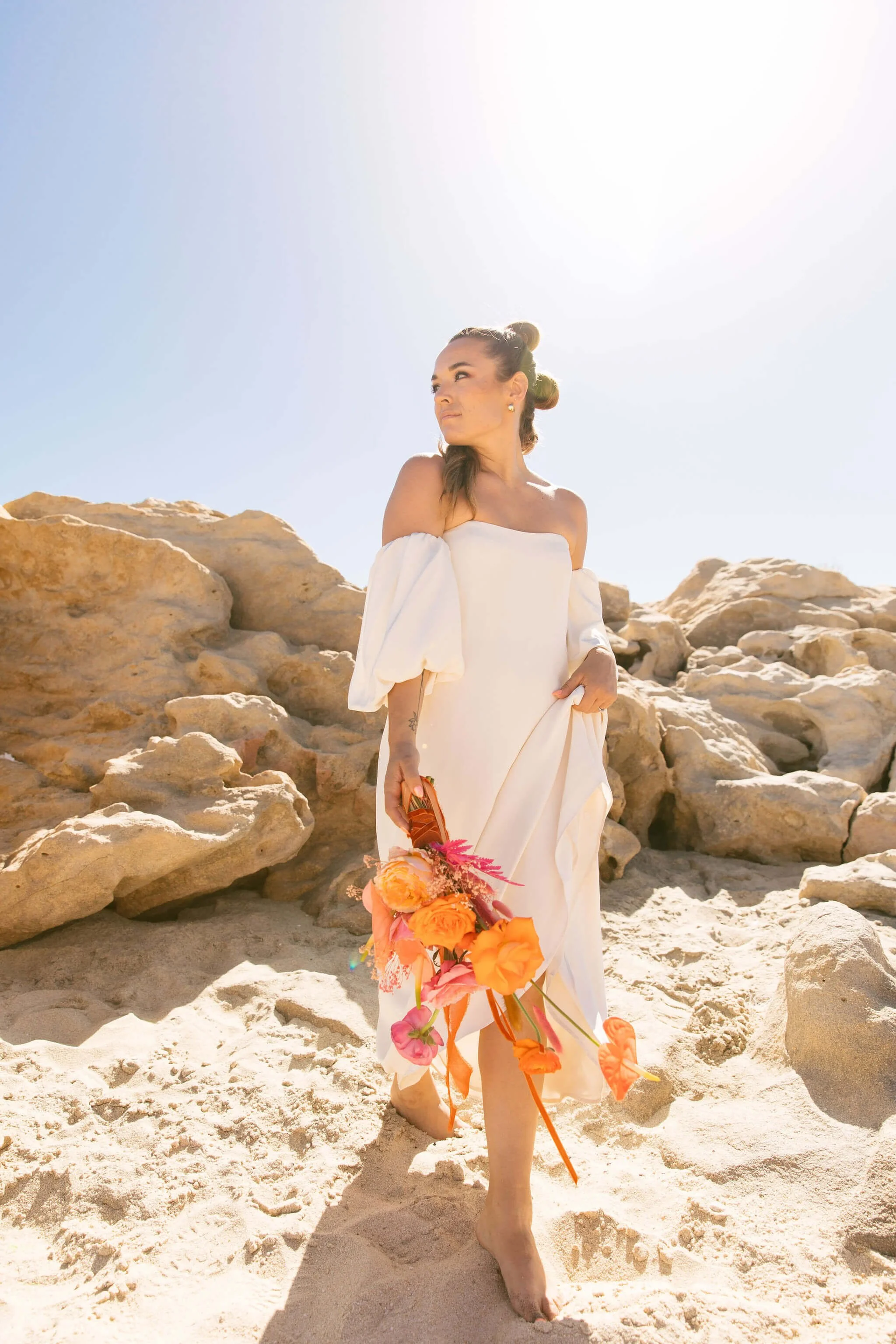 A woman in a white dress holding a colorful bouquet stands barefoot on a sandy beach with rocks around her under a bright, clear sky.
