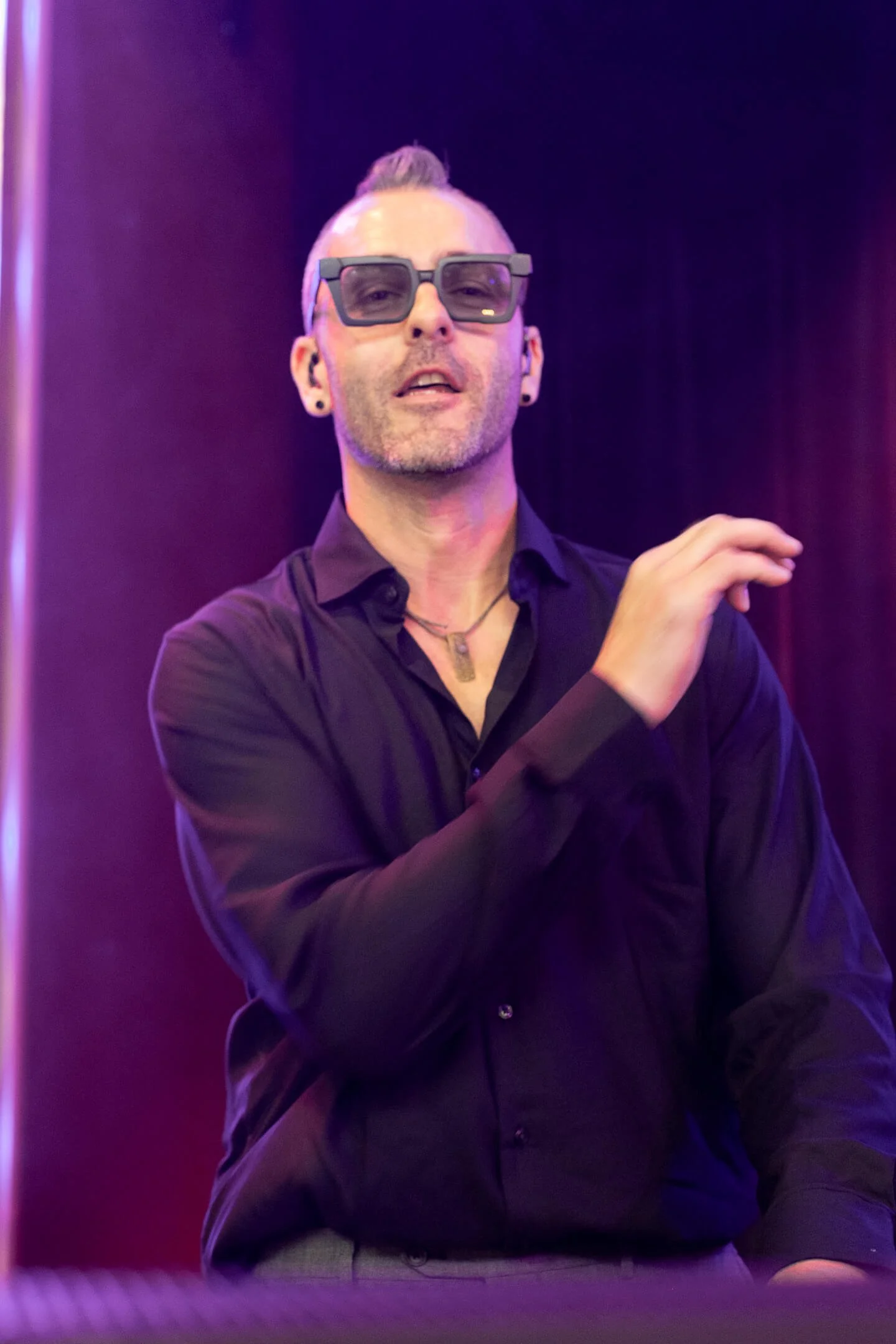 A man with short, dyed platinum blonde hair styled in a mohawk, wearing black sunglasses, earrings, a necklace, and a dark button-up shirt, standing in front of a dark background with purple lighting.