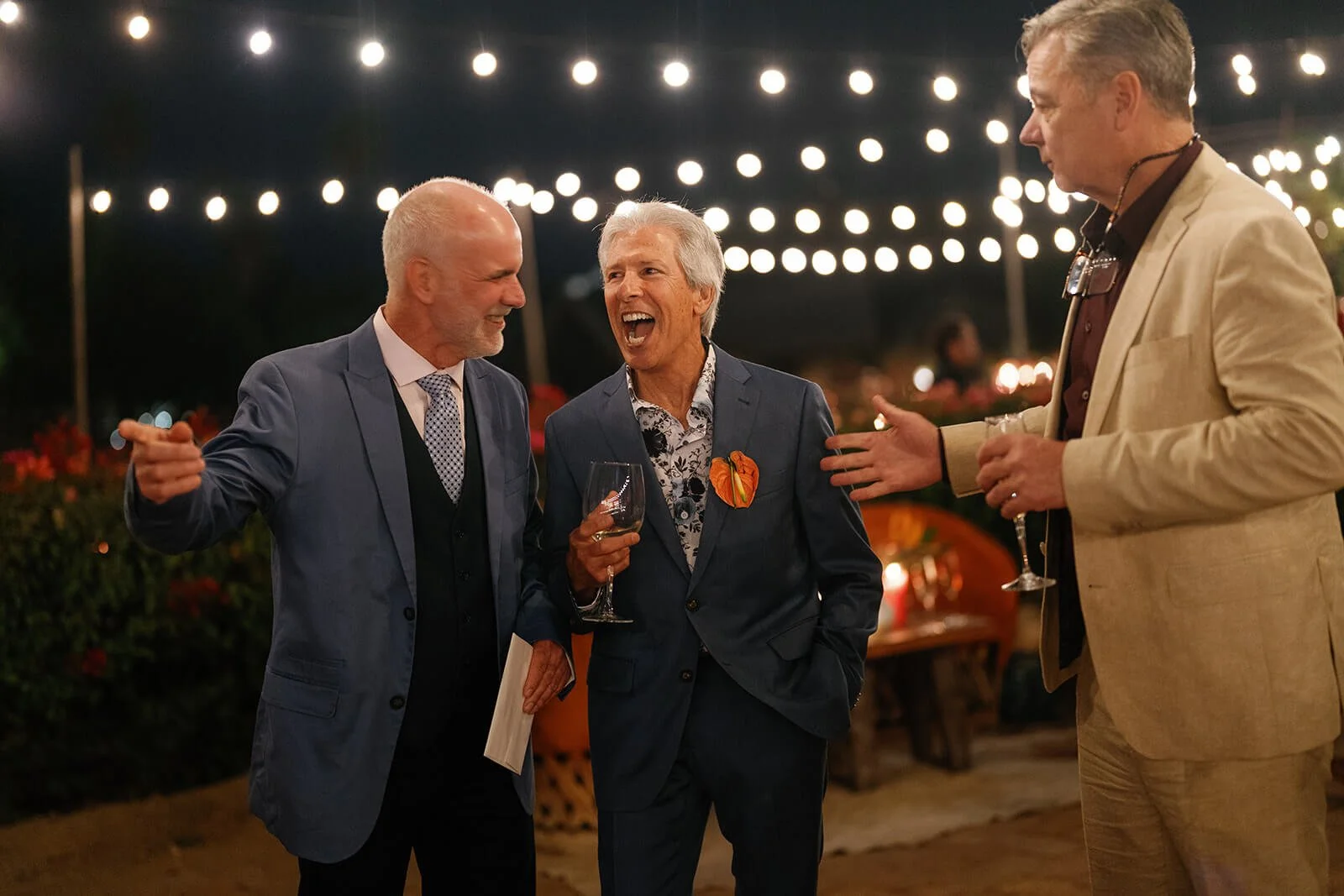 Three middle-aged men in suits at an outdoor evening event under string lights, laughing and conversing while holding glasses of wine.