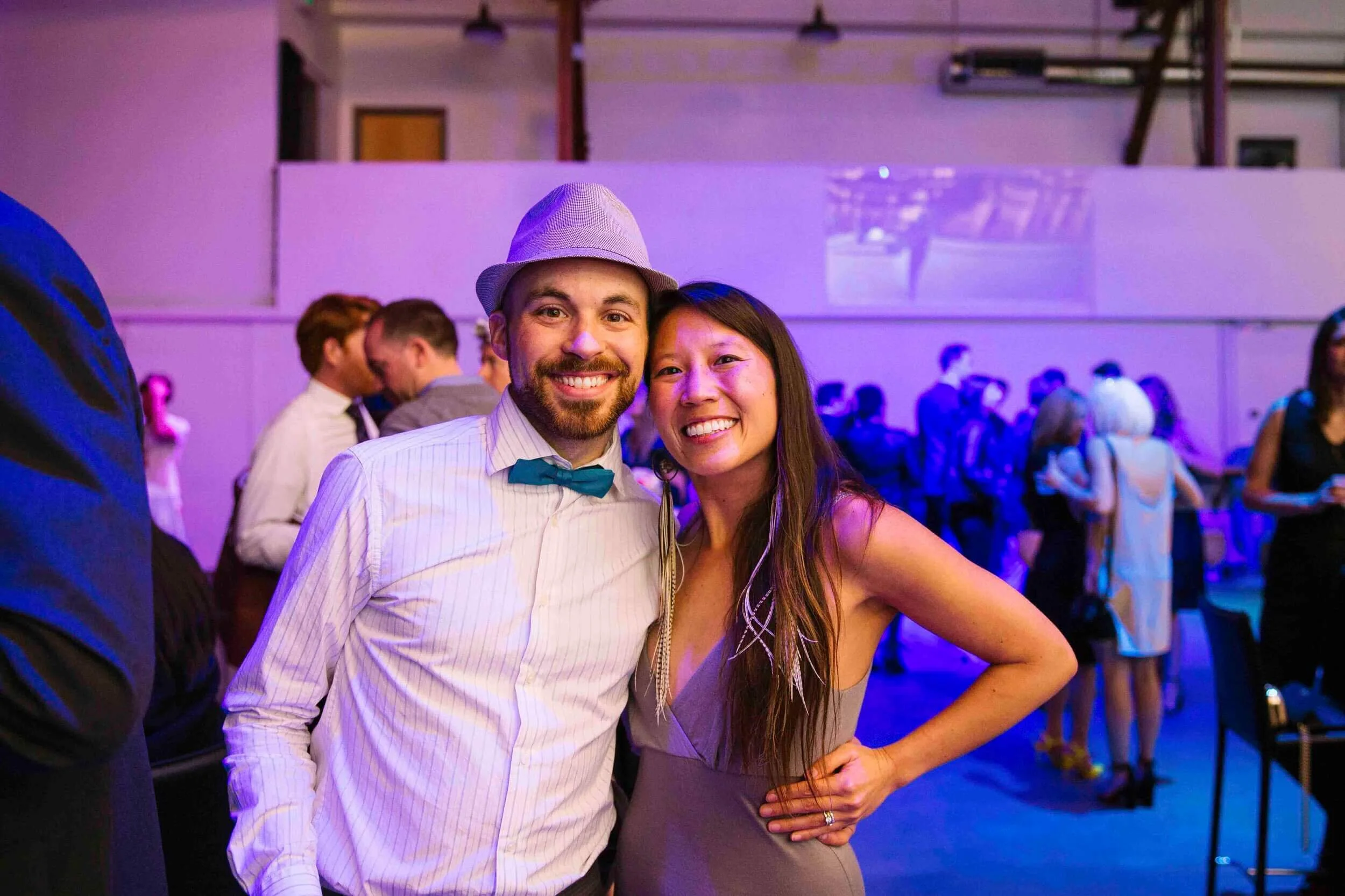 Smiling man and woman at a lively party, standing close together, with the man wearing a light-colored shirt, a bow tie, and a fedora, and the woman wearing a sleeveless dress. The background shows other guests mingling, some dancing, and colorful li