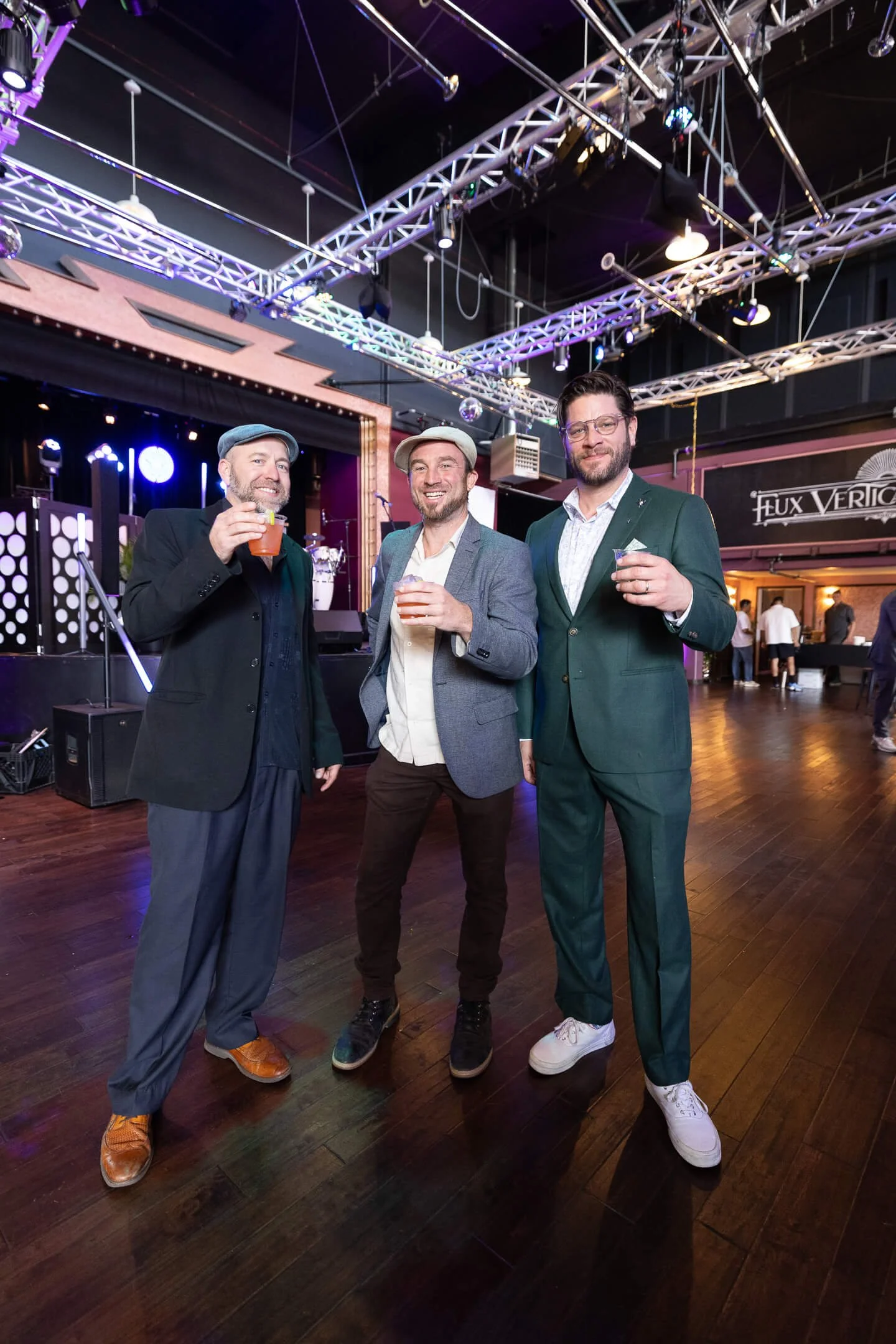 Three men in suits and dress shoes standing in a dimly lit venue with a stage behind them, holding drinks and smiling at the camera.