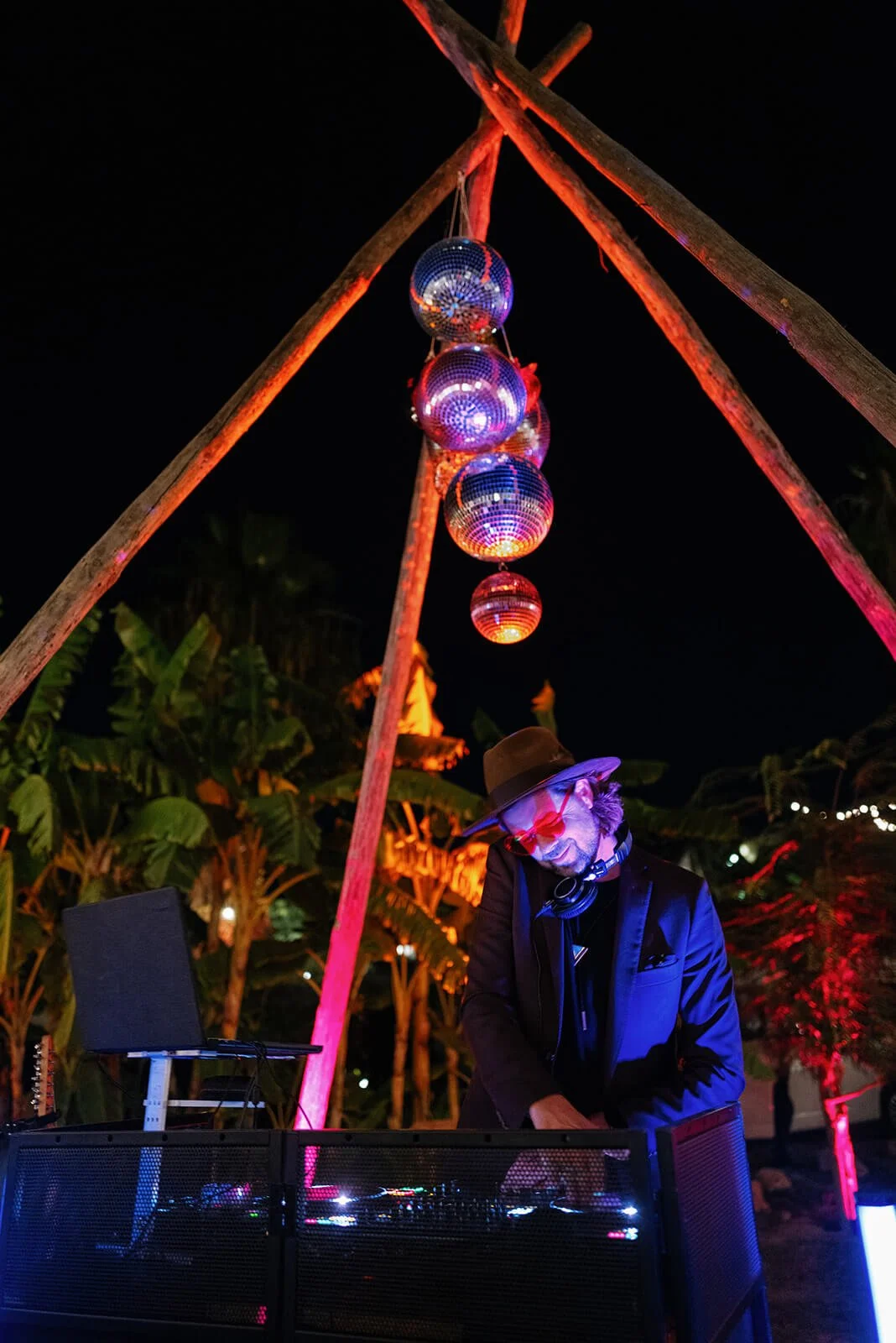 A DJ wearing a brown hat, glasses, and a black suit is performing at an outdoor night event. The scene is lit with colorful lights, and there are five disco balls hanging above the DJ against a dark sky. Wooden poles form a structure around the disco