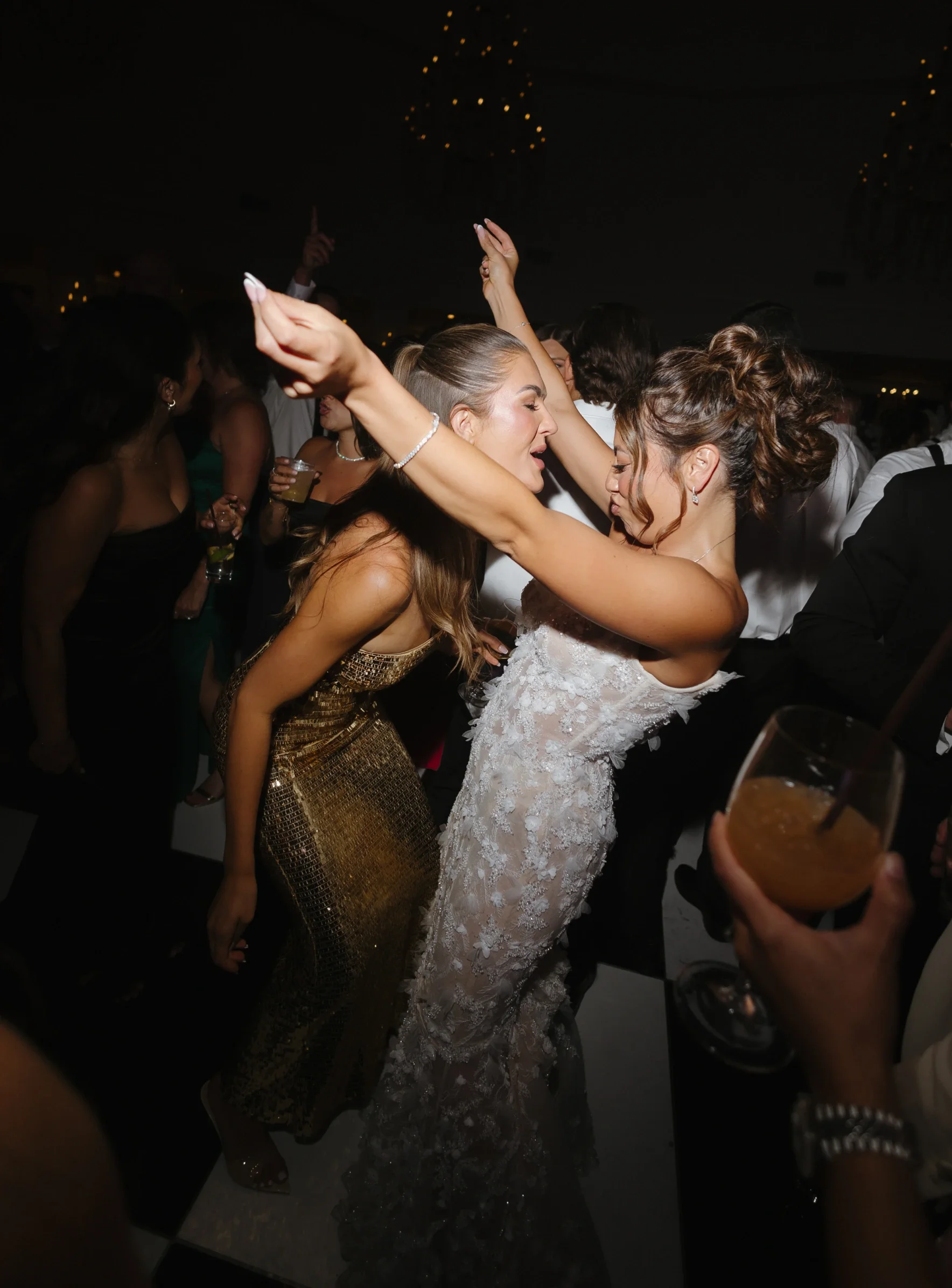 Two women dancing closely at a party, with other people in the background, some holding drinks, under a dark ceiling with string lights.