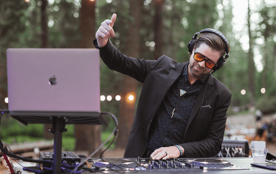 A male DJ wearing headphones and a black blazer mixing music outdoors at sunset with trees in the background, on a wooden platform decorated with flowers, and two pink-lit LED stage lights.