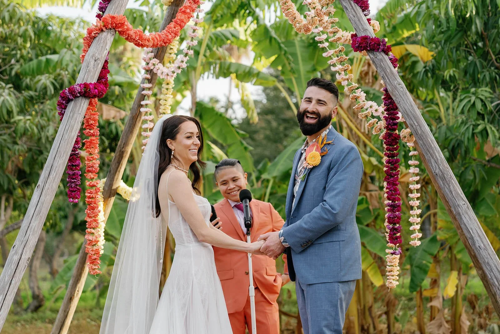 A wedding ceremony outdoors with a couple holding hands and smiling, under a wooden arch decorated with colorful flowers, with a young officiant standing behind them, in a lush green setting.