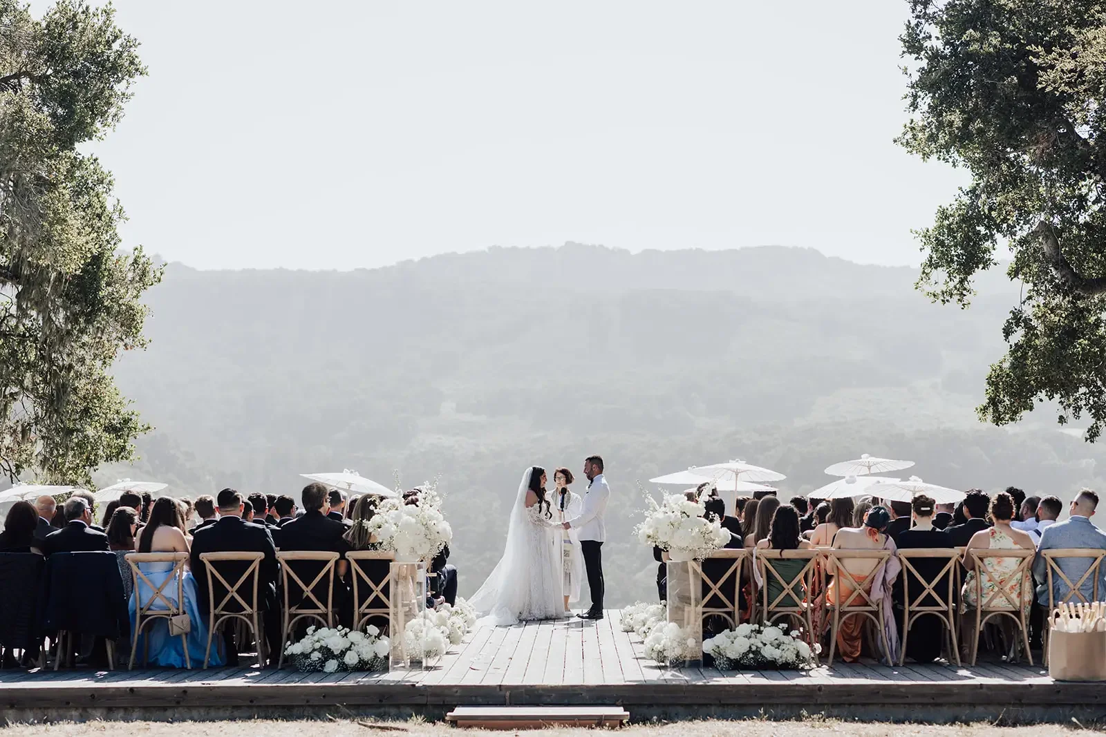 A wedding ceremony taking place outdoors with the bride and groom exchanging vows on a wooden platform, surrounded by guests seated in chairs, with white floral arrangements and umbrellas, against a backdrop of mountains and trees.