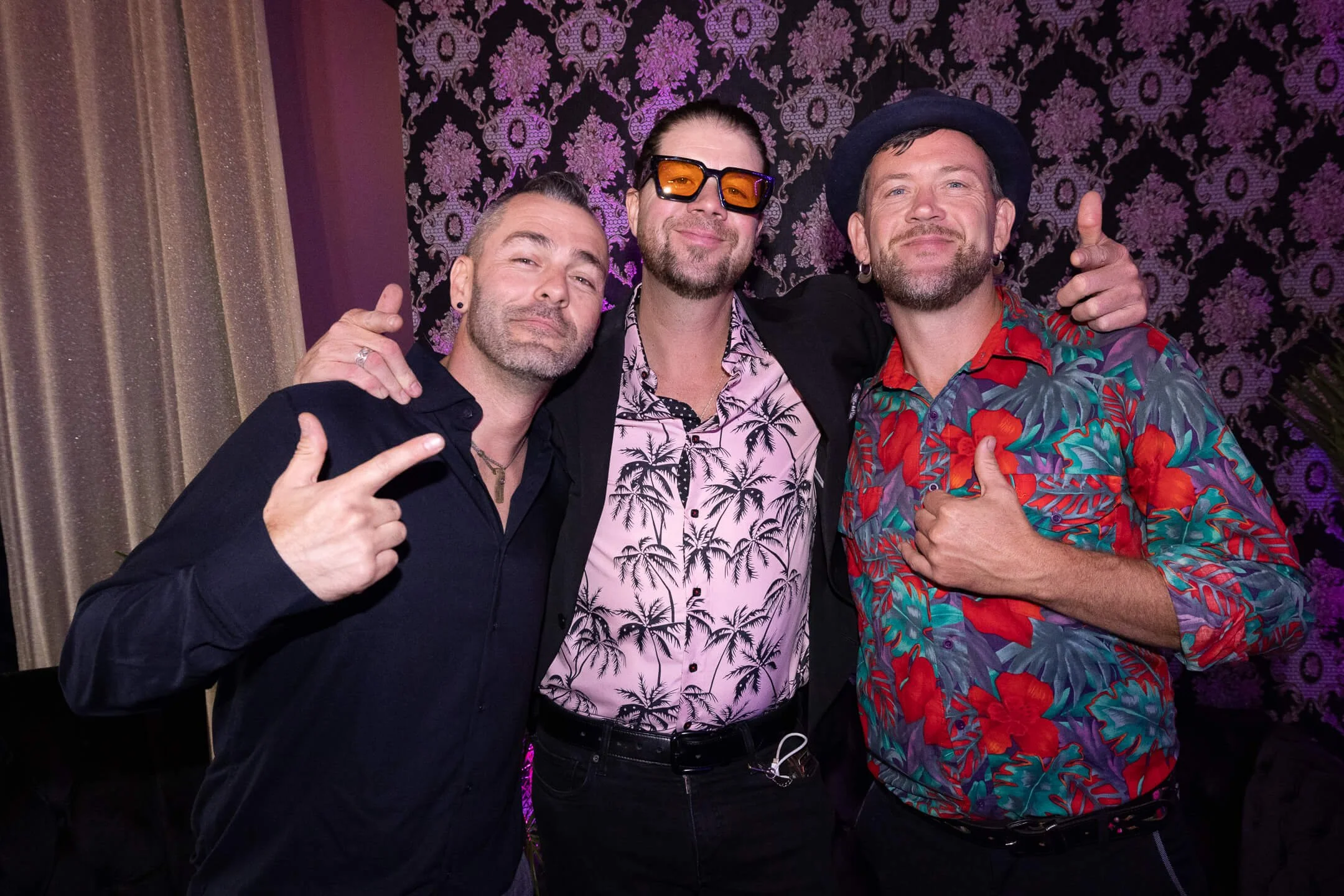 Three men posing together at a party, smiling and showing hand gestures. The man on the left is pointing with his index finger, the man on the right is giving a thumbs-up. They are standing in front of a purple and black patterned wall, with gold and