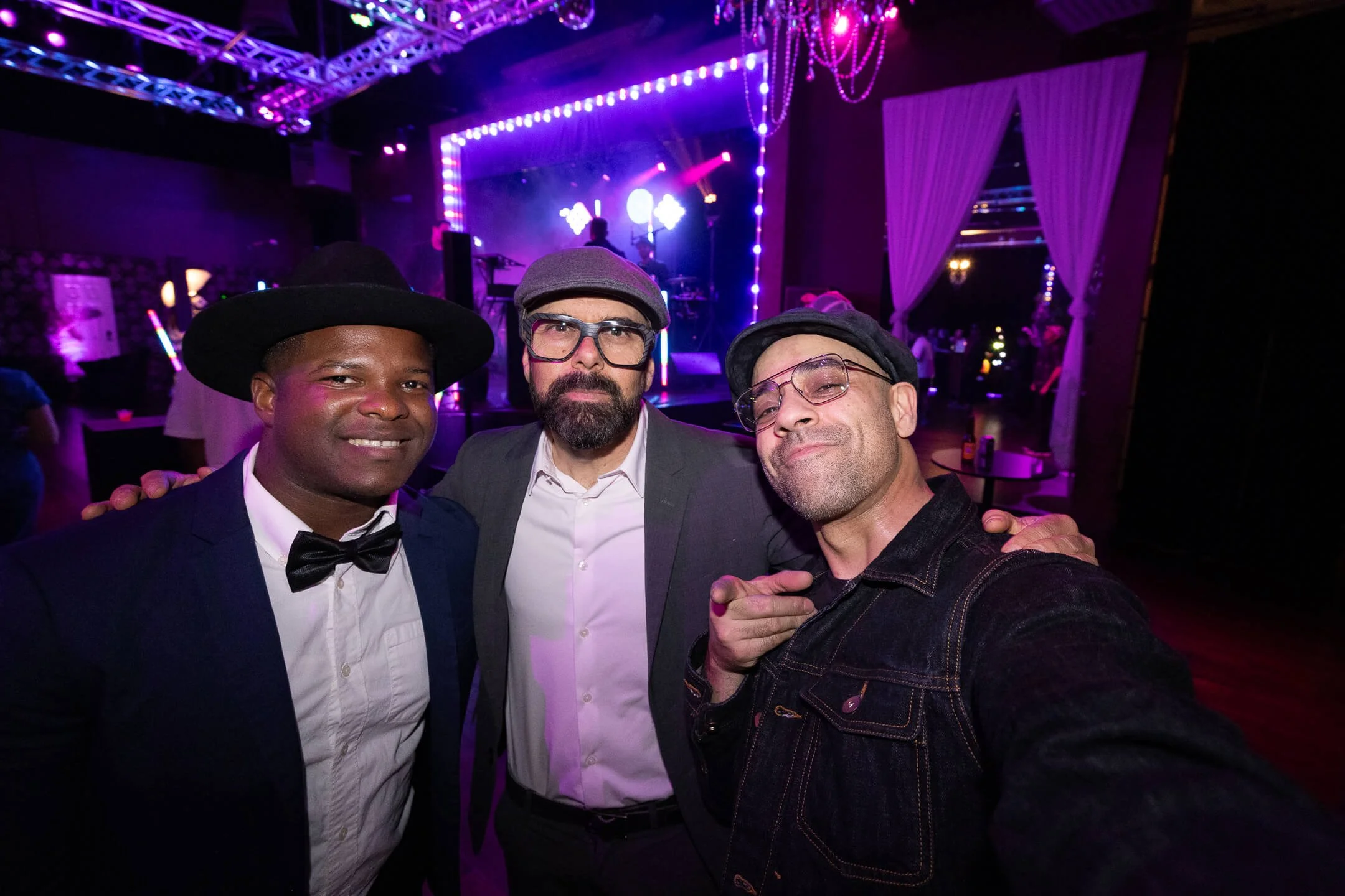 Three men taking a selfie at a party or event with a stage and purple lighting in the background.