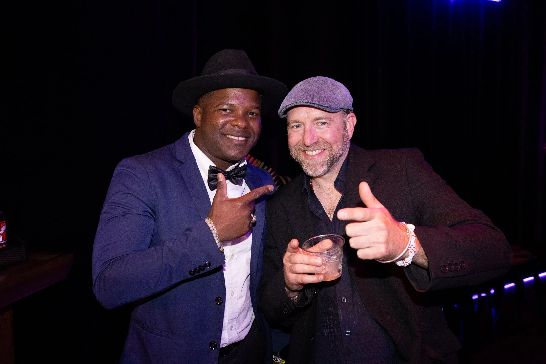 Two men smiling and pointing at each other, one wearing a suit with a bow tie and hat, and the other wearing a blazer with a grey cap, holding a drink, in a dimly lit environment.